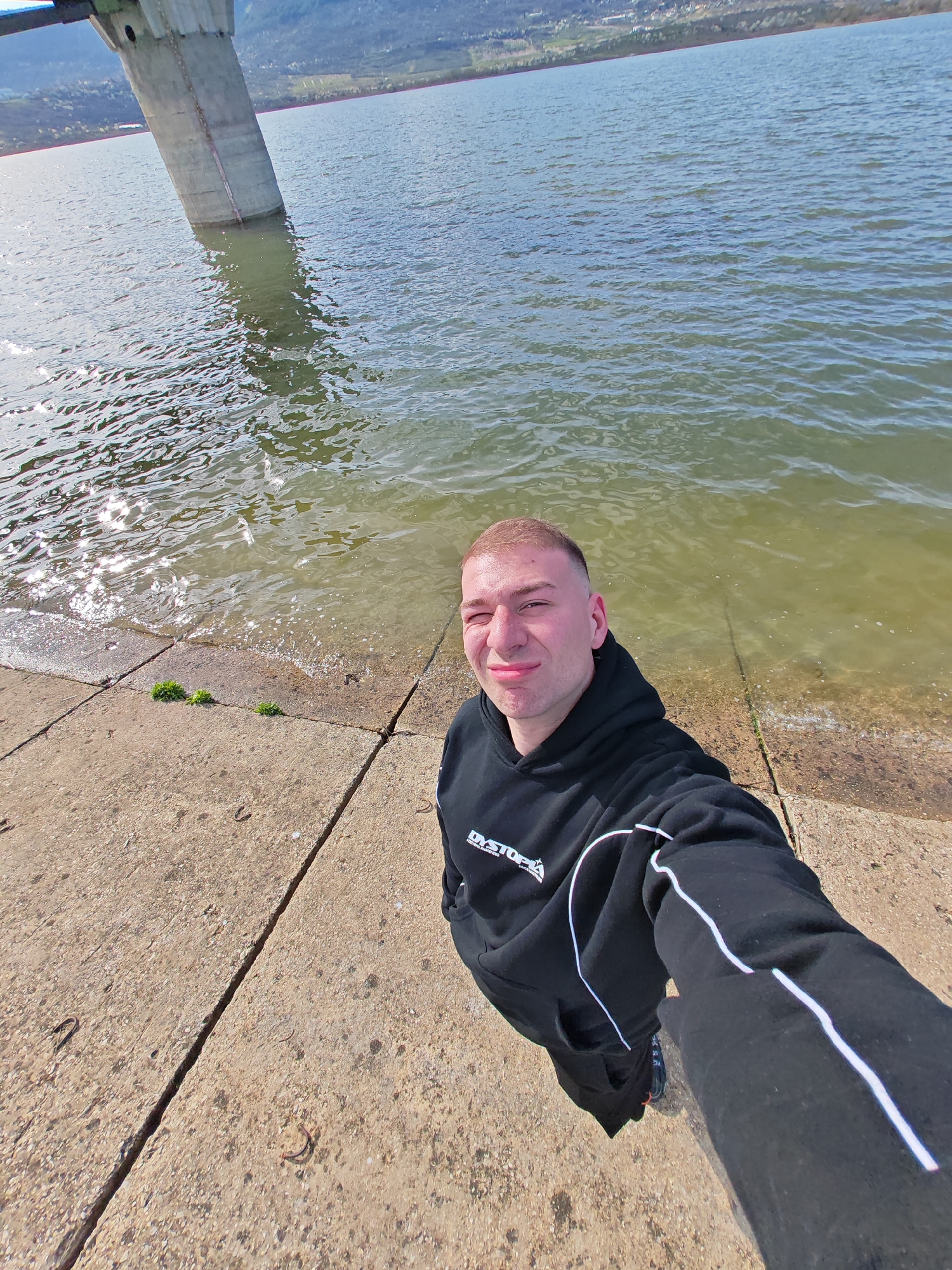 A man is standing on a concrete platform next to a body of water, likely a lake or reservoir, near the city of Shumen, Bulgaria. The man is fair-skinned with short brown hair and is wearing a black hooded sweatshirt and matching sweatpants with a white stripe down the sleeve and pant leg. The hoodie has "DYSTOPIA" printed on the left side. He is holding the camera at arm's length, taking a selfie. Behind him is water, and a concrete support structure is partly submerged in the water. The background includes distant hills or mountains. The weather appears to be sunny and clear.