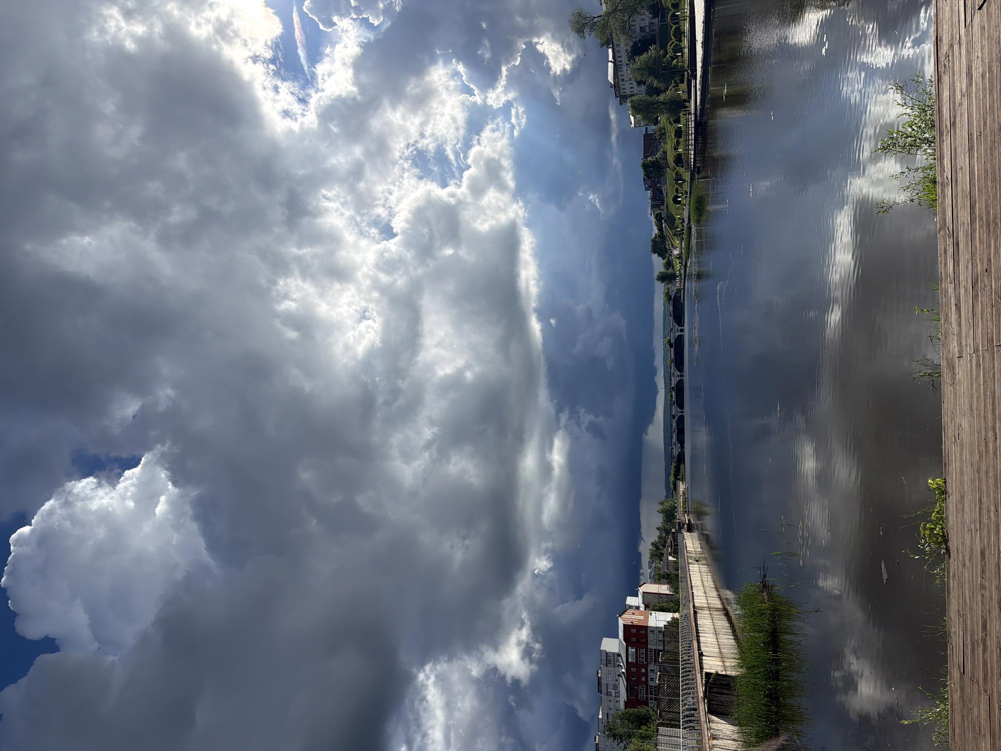 This media file presents a scenic waterfront view in Votkinsk, Russia. The image, originally captured in landscape orientation but displayed rotated 90 degrees clockwise, is dominated by a dramatic sky and a reflective body of water.
The sky is a prominent feature, filled with large, textured clouds. Bright sunlight streams through gaps in the clouds, creating visible crepuscular rays that pierce the atmosphere. Patches of clear blue sky are visible, especially towards the upper right, while other cloud formations appear dark gray and heavy, suggesting dynamic weather conditions, possibly after a recent shower or during intermittent sunshine.
The wide body of water, likely a river or a large lake, occupies the mid-ground. Its surface is relatively calm, yet shows ripples that catch and reflect the light. The water acts as a mirror, clearly reflecting the varied tones of the sky, from brilliant white cloud formations to darker blue and gray areas.
Along the far bank (left side of the un-rotated image), a developed urban or semi-urban area is visible. It features multi-story buildings, possibly residential, nestled among green trees and manicured lawns. A walkway or promenade appears to run parallel to the water. In the distance, a long bridge with multiple arches spans the water, connecting the two sides.
The near bank (right side of the un-rotated image) is bordered by a wooden boardwalk or decking, providing a viewing area along the water's edge. Sparse green vegetation and bushes grow between the boardwalk and the water.
No people or specific activities are visible, lending a sense of tranquility to the scene. The notable details include the striking contrast between the sunlit sky and the darker clouds, indicating changeable weather and suggesting either early morning or late afternoon light. The overall impression is one of natural beauty combined with urban infrastructure, reflecting a serene yet powerful atmosphere. No discernible text is visible within the image.