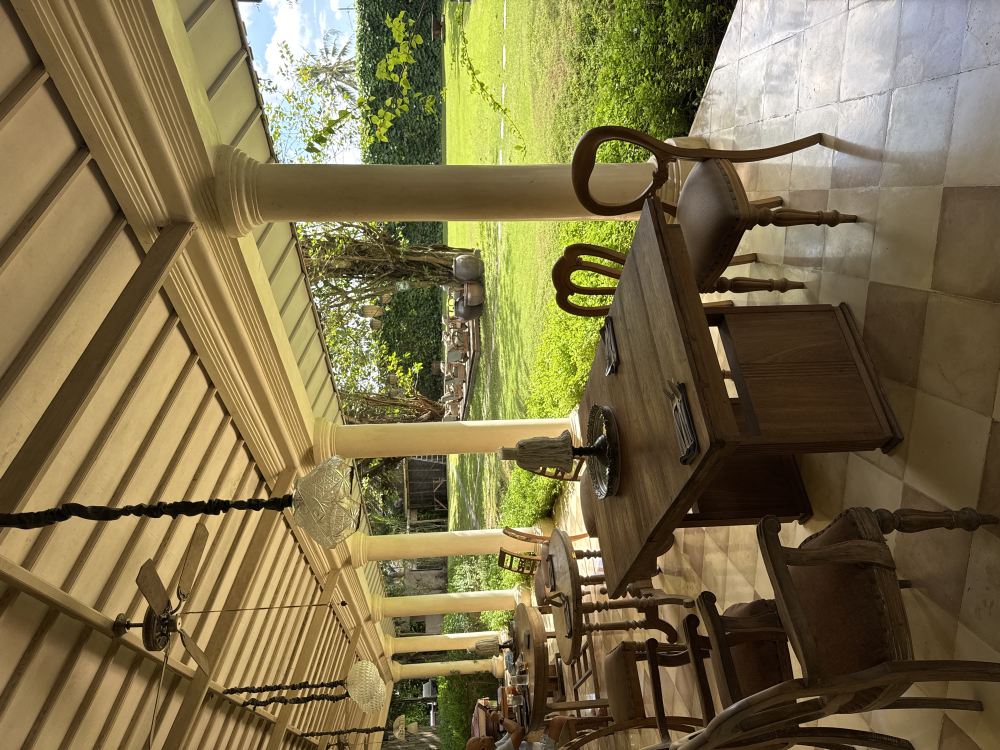 The image depicts an outdoor dining area in Kuta Selatan, Indonesia. The area is covered by a roof supported by several columns. Under the roof, there are tables and chairs arranged for dining. A person is visible in the background, possibly a patron. The dining area overlooks a green lawn with trees and other vegetation. The sky is partially visible in the background.