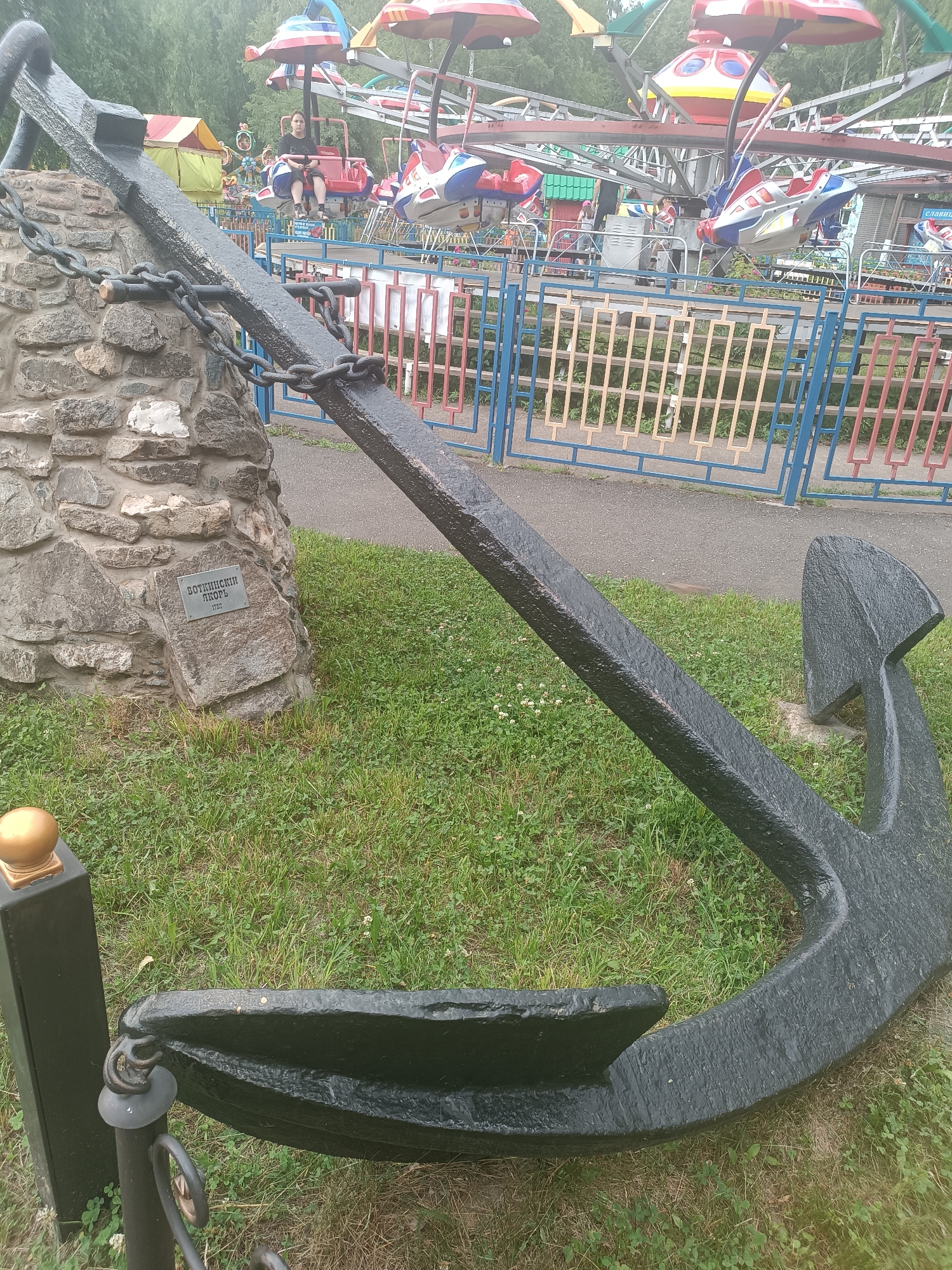 The image shows an outdoor scene at an amusement park in Chaikovsky, Russia. In the foreground, a large, black metal anchor rests on grass, secured to a stone monument by thick chains. A plaque on the stone monument reads "Воткинский якорь 1937" (Votkinsk Anchor 1937). To the left of the anchor, a black metal post with a golden ball on top is visible. In the background, a children's amusement ride with colorful cars, some shaped like airplanes, is in operation. Several people are seated in the ride cars. A blue and red fence separates the ride area from the foreground. The environment is green with grass and trees visible in the distance.