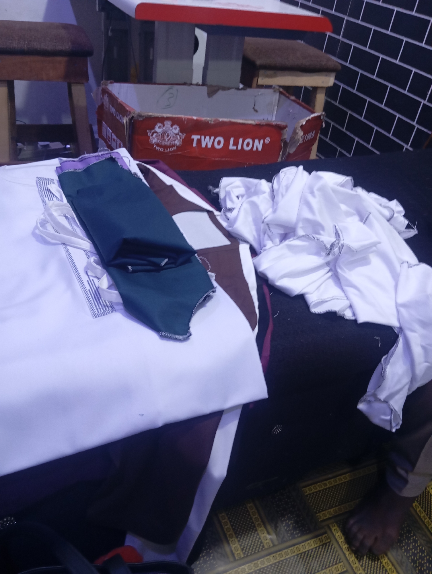 The image captures an indoor scene with several fabric items laid out on a dark surface, possibly a table or floor. In the foreground, a white fabric with a patterned border is visible, partially covered by a folded piece of dark green fabric. To the right, a pile of crumpled white fabric with ruffled edges is present. In the background, a red and white cardboard box with "TWO LION" branding is partially visible, along with two wooden stools. The lighting appears to be artificial, creating a somewhat dim atmosphere. The overall scene suggests a crafting or tailoring area, with fabric and related materials.