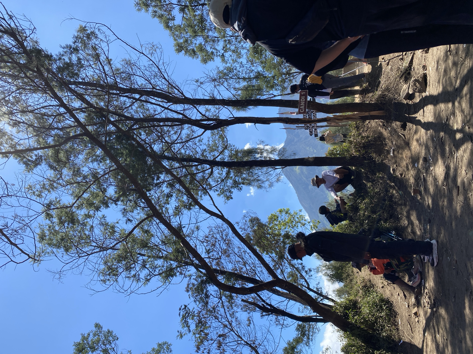 A group of people are gathered on a dirt path near a mountain in a dry, sunny environment. Trees with sparse foliage and thin branches dominate the foreground and mid-ground. In the background, a conical mountain is visible under a clear blue sky with scattered white clouds. Several individuals are visible in the scene. One person in the foreground is wearing a black jacket and a black cap, facing away from the camera. Another person, further back, wears a light-colored hat and a white shirt, sitting on the ground. To their left, someone in an orange jacket and dark pants is seated. In the mid-ground, a signpost with the text "CANDI 1" is visible. Two people stand near the signpost, one in a dark shirt and the other in a lighter colored top. Other individuals are dispersed throughout the scene, some appearing to be resting or observing the surroundings. The ground is covered in dirt, small rocks, and sparse vegetation. Shadows from the trees are cast across the path. The overall impression is of a hiking or trekking location.