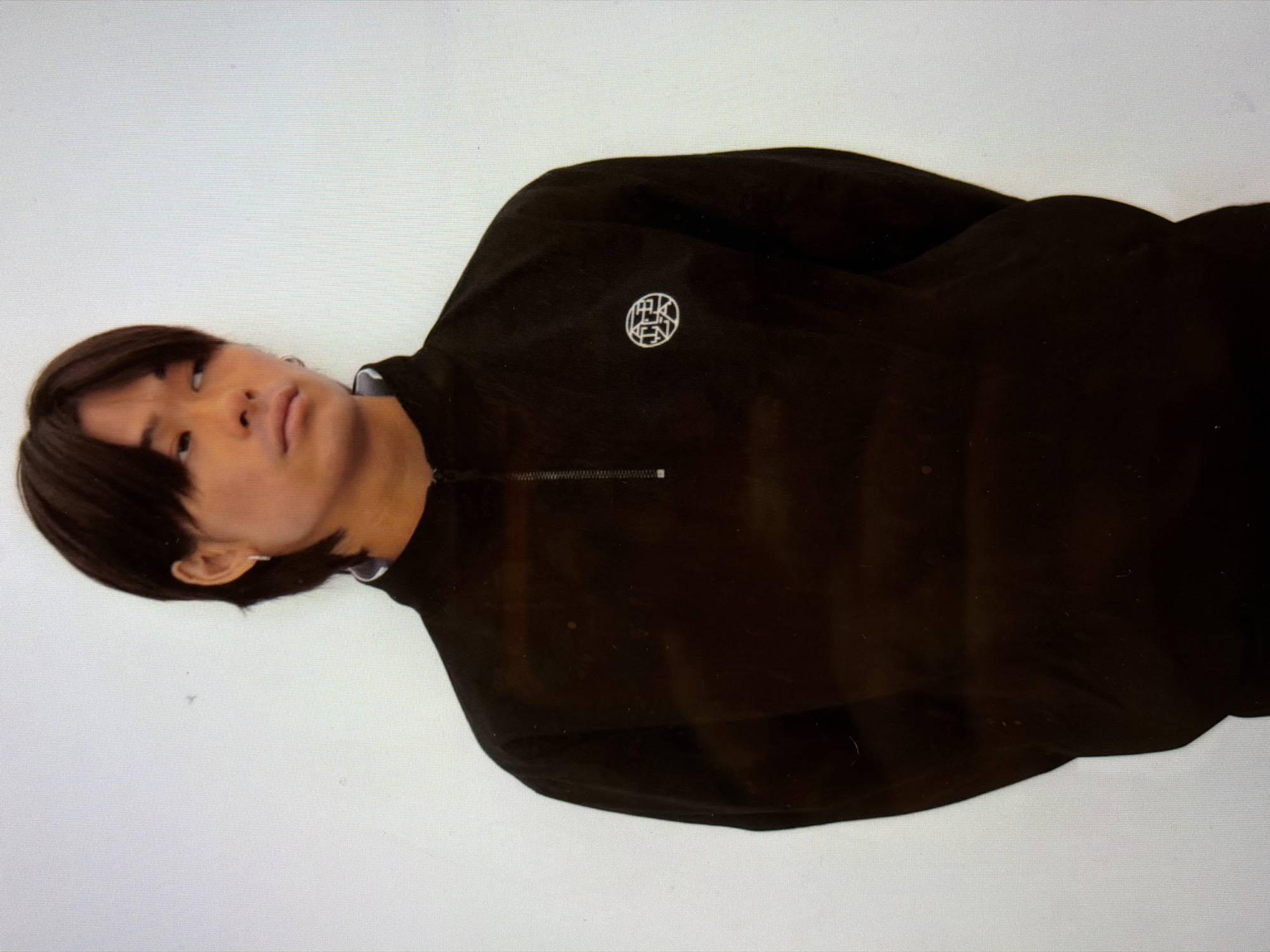 A person is laying down on a white surface. The person has short dark hair and is wearing a black shirt with a zipper at the neck and a logo on the left chest. The person is looking up and to the left. The location is unknown, but it is not possible to determine if the image was taken in New York City.