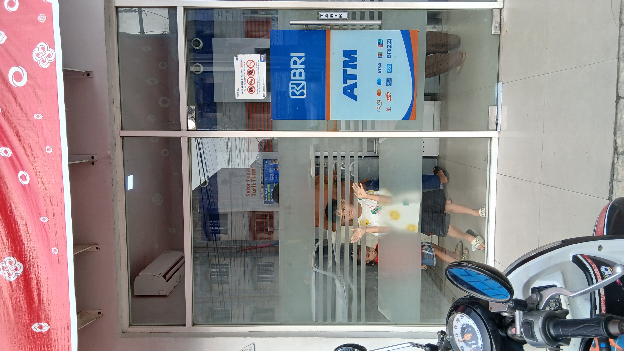 This image depicts the entrance to a BRI (Bank Rakyat Indonesia) ATM facility in the City of Medan, Indonesia. The entrance features large glass doors with various signage. On the right door, a prominent blue and orange sign indicates "ATM" with the BRI logo at the top. Above this, a smaller white sign with red text displays rules and regulations, including a prohibition against smoking and a reminder to maintain a safe distance.
Through the glass, two children are visible inside the facility. A boy in a white t-shirt with yellow smiley faces stands with his hands raised, facing the camera. To his left, a younger child in a red shirt is partially visible. The interior of the ATM area is seen through the glass, with reflections showing a white air conditioning unit mounted on the wall and some interior architectural details.
Outside the entrance, a motorcycle is partially visible in the foreground on the left side of the image, with its handlebars and a side mirror in view. The ground outside the entrance is tiled. The overall scene suggests an active, public space associated with banking services.