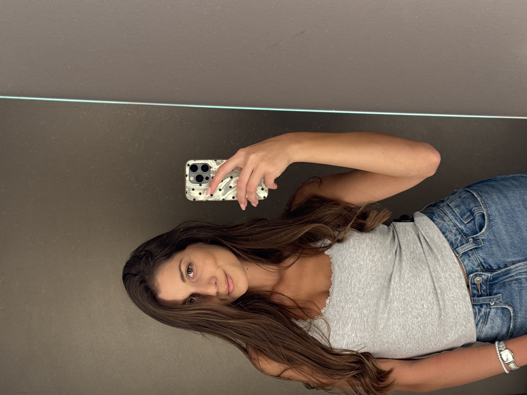 A woman is taking a selfie in a mirror. She is wearing a gray tank top and blue jeans, and has a silver watch on her wrist. She is holding a phone with a black and white polka dot case. The background is a dark gray wall with a thin white line running horizontally across the top. There is no indication of the city of Burgas, Bulgaria.