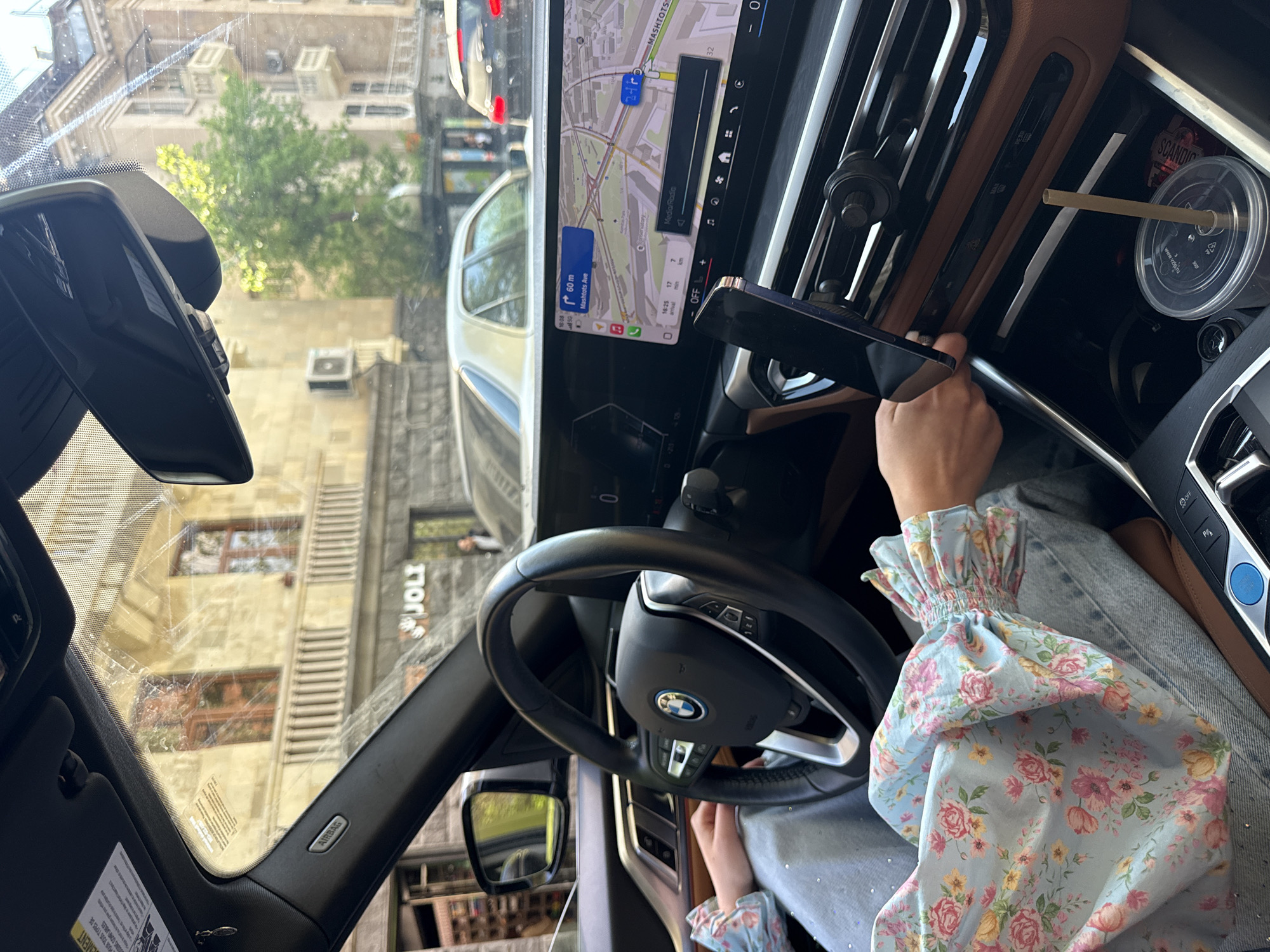 The image captures a driver's perspective from inside a modern car, looking out onto a city street during daytime. The primary subject is the car's interior, featuring a prominent dashboard with a large infotainment screen displaying a navigation map. A person's right arm and hand are visible, wearing a light blue top with a floral pattern of pink and yellow roses, with a ruffled sleeve cuff. The hand appears to be near a phone mounted to the dashboard, or interacting with car controls.
Notable objects inside the car include a black BMW steering wheel with the brand's logo, a rearview mirror, and an "AIRBAG" label on the A-pillar. To the right, a clear plastic cup with a beige straw and a "SCANDIC" logo is visible, positioned near the air vents and other controls. The car's interior also features brown trim accents.
The navigation screen shows a map centered around "MASHTOTS" and indicating "Mashtots Ave" with a distance of "60m," and "Conservatory." The time "16:25" and an estimated travel time of "17 min" are also displayed on the screen. The visible text "Media/Radio" suggests entertainment options.
Outside the car, through the windshield, several multi-story buildings are visible, typical of an urban environment. A green tree stands between buildings. One building clearly displays a sign with "JOLI." The bright lighting suggests it is sunny. Given the navigation details showing "Mashtots Ave" and the provided context, the location is identified as Yerevan, Armenia.
