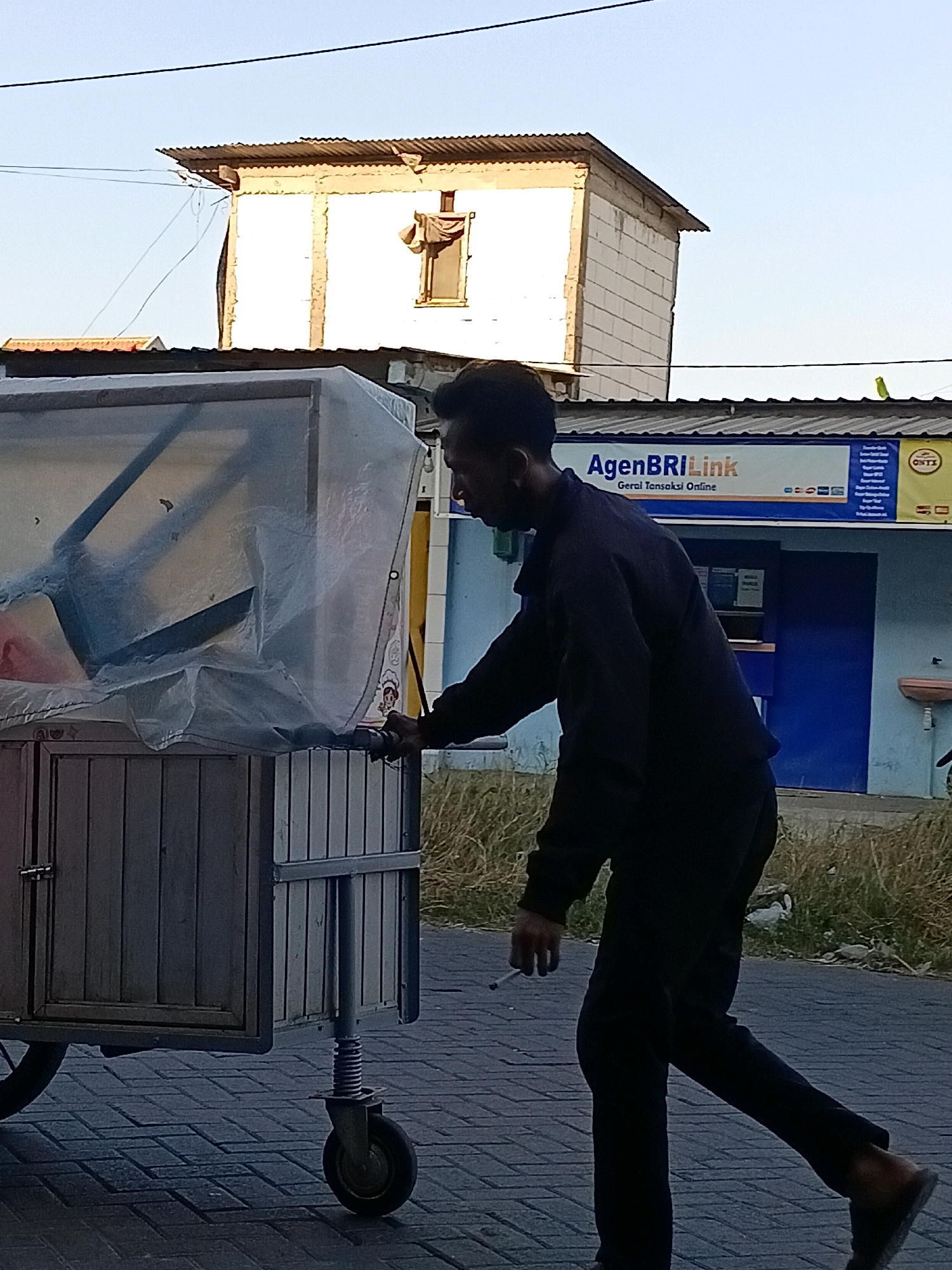 A man is pushing a cart with a clear plastic cover on a paved sidewalk in Kletek, Indonesia. The man is wearing a dark long-sleeved shirt and dark pants, and he is holding a cigarette in his right hand. The cart is made of wood and metal, and it has a spring-loaded wheel in the front. In the background, there is a building with a sign that says "AgenBRI Link". The building is blue and white, and it has a window. The sky is clear and blue. The environment appears to be a street in a residential or commercial area.