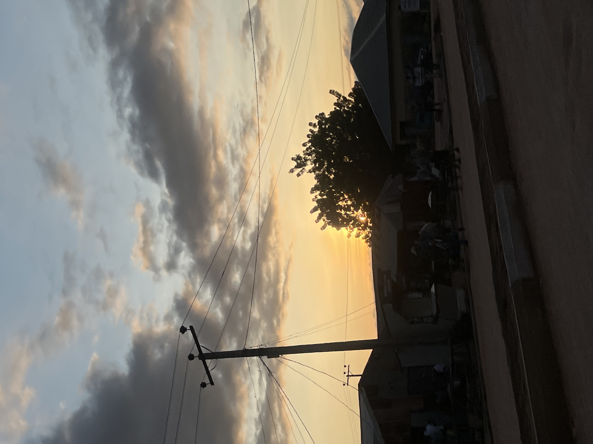 The image depicts an outdoor street scene in Howayi, Nigeria, captured during either sunrise or sunset, characterized by strong backlighting from a low sun on the right horizon. The sky transitions from dark grey, dense clouds in the upper left to a pale blue with scattered, peach-tinted clouds in the upper right, before becoming a vibrant golden-yellow towards the horizon. A prominent, dark utility pole stands left of center, silhouetted against the bright sky, with multiple power lines extending horizontally and diagonally across the frame.
To the left, a dark, single-story building with a sloped roof is visible. In the mid-ground, a series of dark, indistinct structures, possibly shops or stalls, are present, some with visible awnings or umbrellas. On the right, a larger building with a grey, sloped roof and lighter-colored walls is largely obscured by shadow. A dense, dark-foliaged tree on the right partially obscures the bright sun, which casts a strong golden glow through its leaves.
The foreground consists of a dirt or unpaved road, bordered by concrete curbs or low barriers that run horizontally across the lower portion of the image. Several human figures, mostly indistinct and silhouetted by the backlighting, are present on the road and near the structures in the mid-ground. They appear to be walking or standing, with some clustered together, but specific actions or interactions are not clearly discernible. The overall scene is heavily shadowed in the foreground and mid-ground due to the intense backlighting, creating a strong silhouette effect against the bright, colored sky.