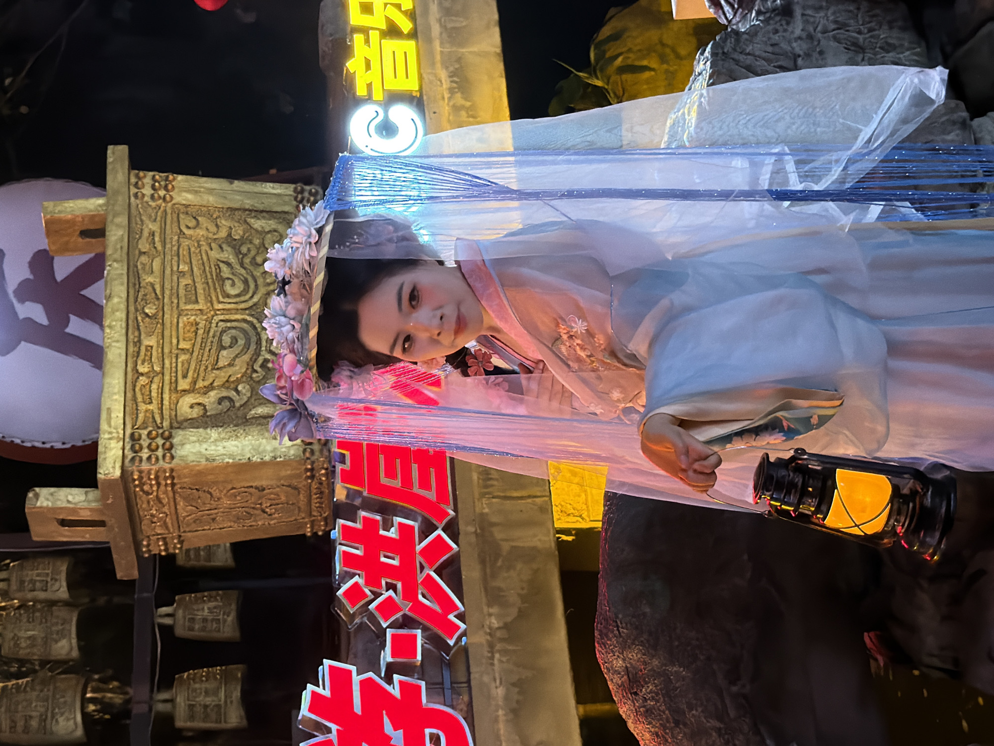 The image captures a scene in Yuzhong District, China, at night. The main subject is a woman adorned in traditional Chinese attire, wearing a delicate veil adorned with flowers. She is posed within a decorative, possibly ceremonial, structure. The setting appears to be outdoors, indicated by the dark background and artificial lighting.
The woman is holding a vintage-style lantern that emits a warm, yellow glow, suggesting evening or nighttime. Her makeup is noticeable, with a focus on her eyes and cheeks, giving her a somewhat ethereal or festive appearance. She is looking towards the viewer with a gentle expression.
Behind her, a large, ornate golden structure is visible, possibly part of a temple or a float for a parade. The structure features intricate carvings and is topped with what appear to be small bells. There are also prominent red neon signs with Chinese characters, adding to the vibrant and possibly celebratory atmosphere. The veil over the woman is made of sheer, shimmering fabric with blue beaded accents.
The overall impression is one of a cultural event or performance taking place in an urban, possibly historical, setting at night. The lighting, attire, and decorative elements all contribute to a visually rich and atmospheric scene. The visible text on the neon signs are Chinese characters, but they are not legible enough to be transcribed.