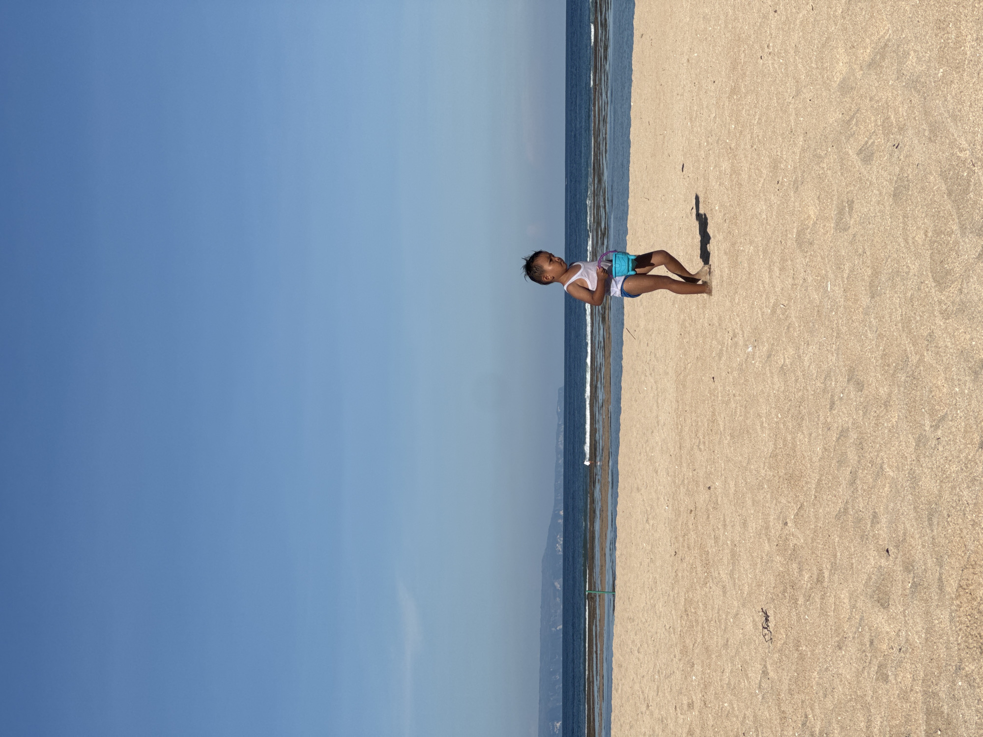The image shows a child standing on a sandy beach next to the ocean in Kuta Selatan, Indonesia. The child is wearing a white tank top and blue shorts and is holding a blue bucket. The sky is clear blue, and the sand is tan. The ocean is visible along the horizon line, with small waves breaking on the shore. A shadow cast by the child is visible on the sand.
