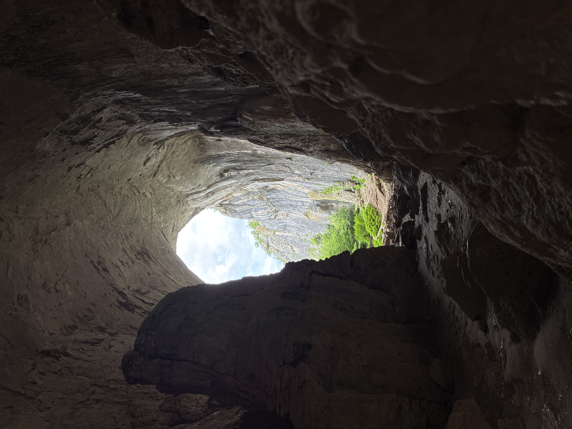 This image shows a view from inside Devetashka Cave in Karlukovo, Bulgaria. The viewpoint is from the interior of the cave, looking up and outward through a large natural opening. The foreground and the surrounding cave walls are composed of rough, dark rock formations. Outside the cave entrance, there is a glimpse of vegetation, some layered rock formations, and a patch of blue sky with white clouds.