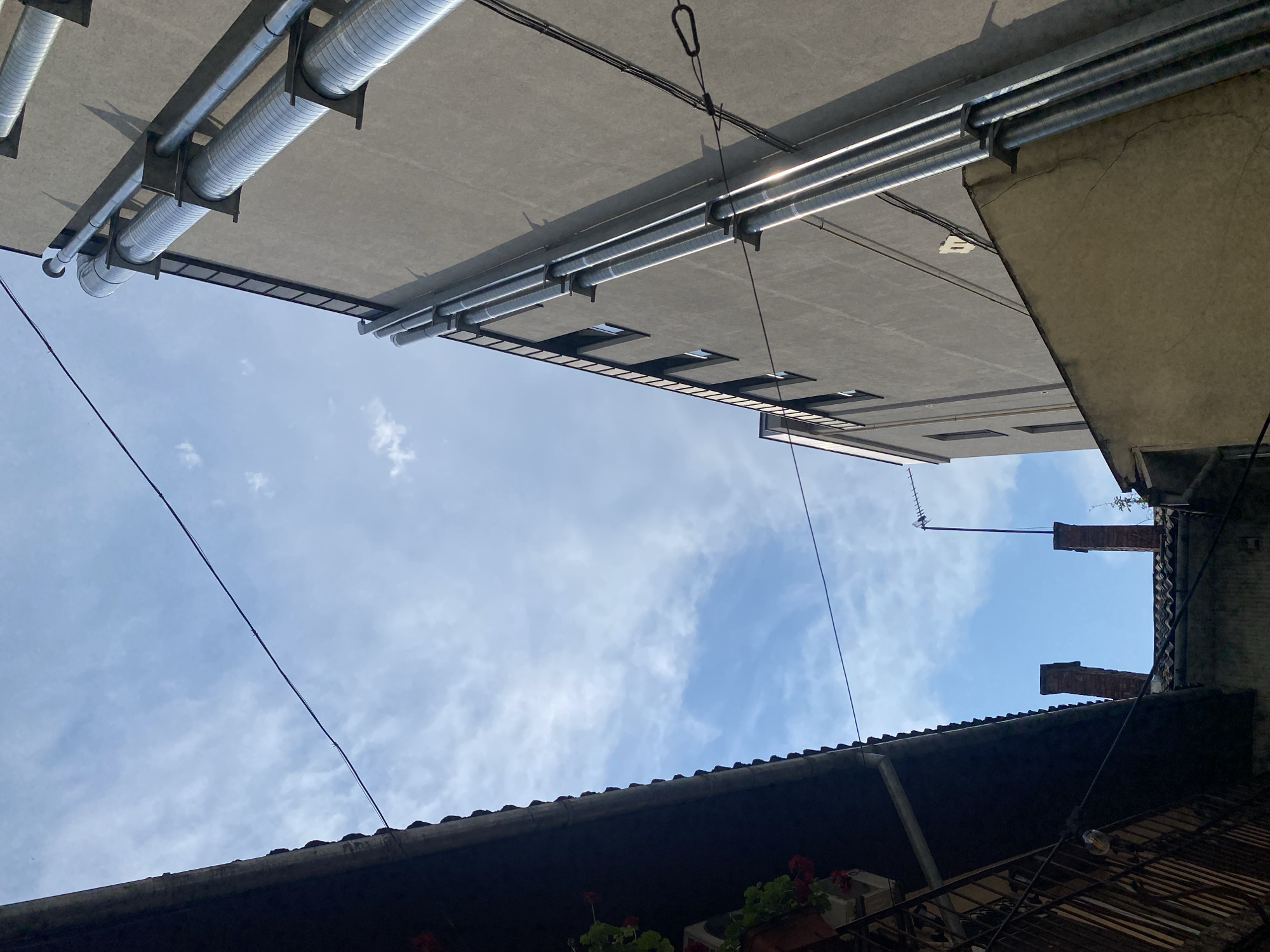 An eye-level, medium shot captures a view looking upwards at buildings and sky. On the left, a dark, tiled overhang with a black metal railing extends diagonally across the frame. Red flowers in pots are visible against the railing. To the right, a tall, gray building features multiple large, corrugated metal pipes running vertically along its side, attached by metal brackets. Several small, dark windows are visible on the gray building. In the distance, two brick chimneys rise above a tiled roof, and an antenna with horizontal elements is mounted on a pole. The sky is blue with scattered white clouds. A thin black wire stretches across the top of the frame.