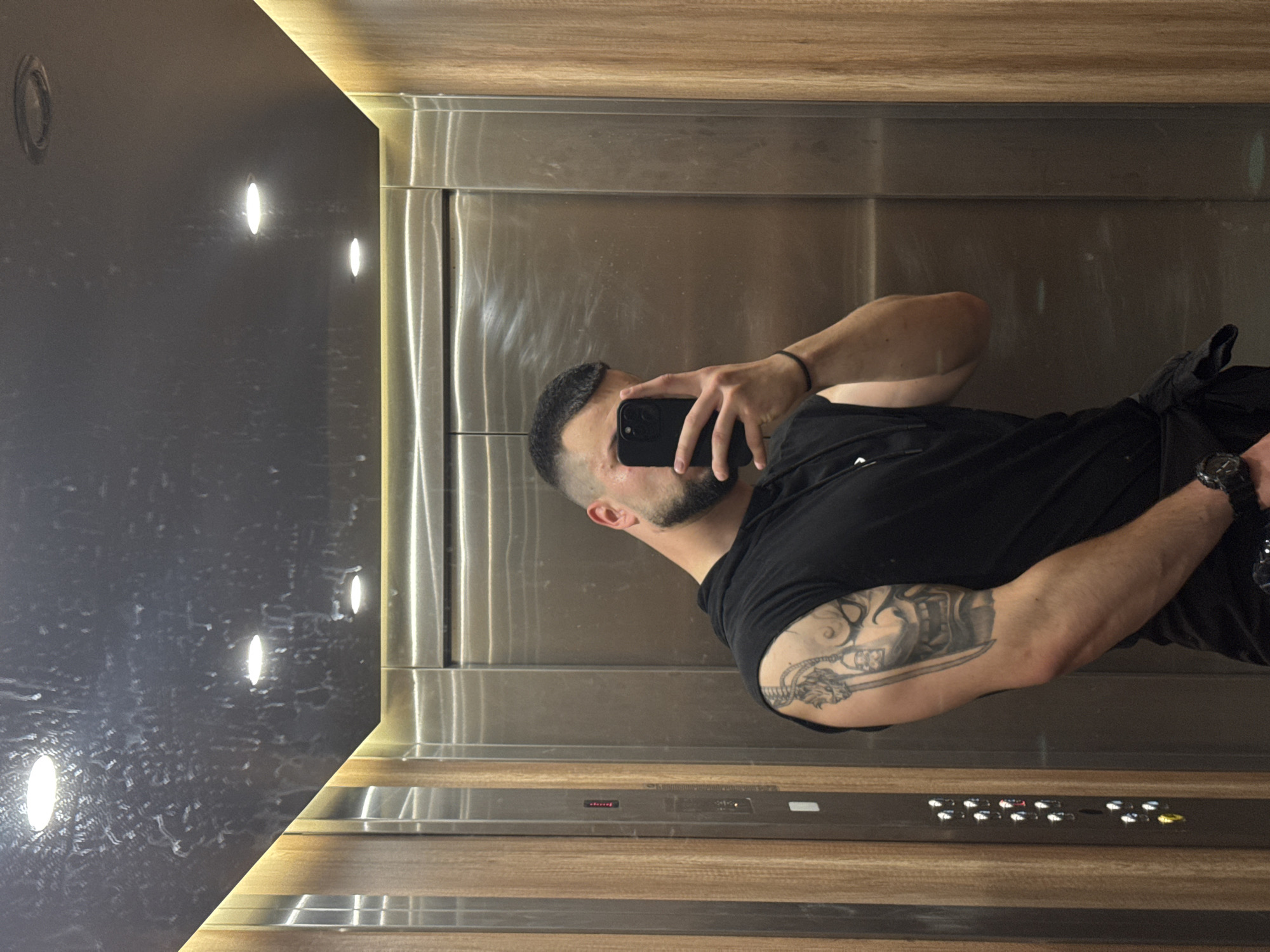 The image, rotated 90 degrees clockwise, depicts a man taking a selfie inside a modern elevator. The man is reflected in the polished metallic surface of the elevator doors. He has a dark beard and short, dark hair with a fade. He is wearing a black short-sleeved t-shirt, a black band on one wrist, and a smartwatch on the other. A large, intricate tattoo featuring a warrior-like figure and a weapon is visible on his left upper arm. He holds a black smartphone up to capture the reflection.
The elevator interior showcases a contemporary design. The left wall is a dark, possibly black or dark grey, textured panel embedded with multiple bright, circular recessed lights. The ceiling and floor sections of the elevator are lined with warm-toned wooden panels, which also feature strip lighting along their edges, contributing to a well-lit ambiance. The right side of the image is dominated by shiny, metallic (likely stainless steel) elevator doors, which create a strong reflection of the man and the internal lighting. Below the doors, an elevator control panel is visible, equipped with several light-up buttons and a small, red-lit digital display, though no specific text or numbers are clearly legible. This modern elevator is located in Sofia, Bulgaria.