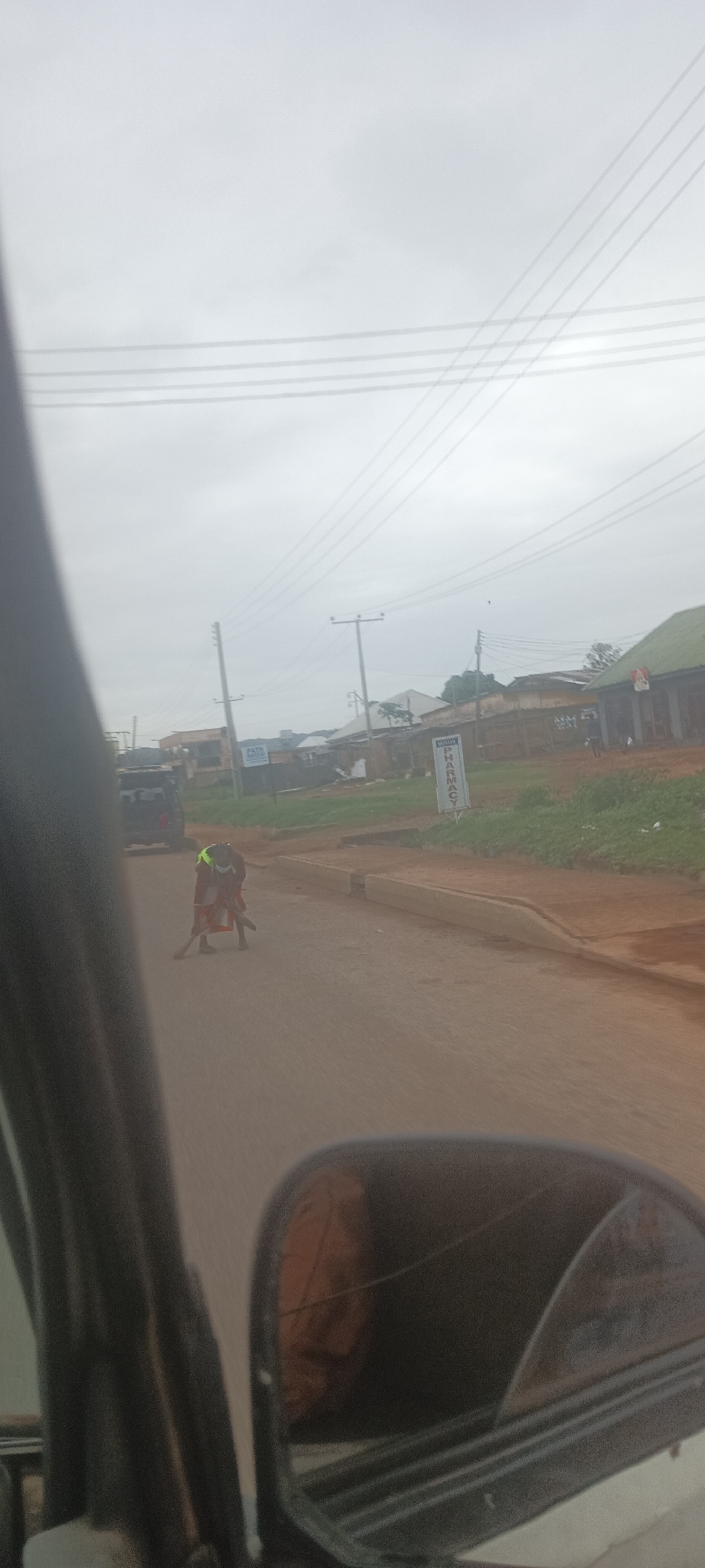 The image shows a road in Bukuru, Nigeria. A person wearing a neon yellow vest and dark clothing is sweeping the road. There are buildings on both sides of the road, and power lines run overhead. A sign for a pharmacy is visible. The photo was taken from inside a vehicle, with a portion of the vehicle visible in the foreground. The weather appears overcast.