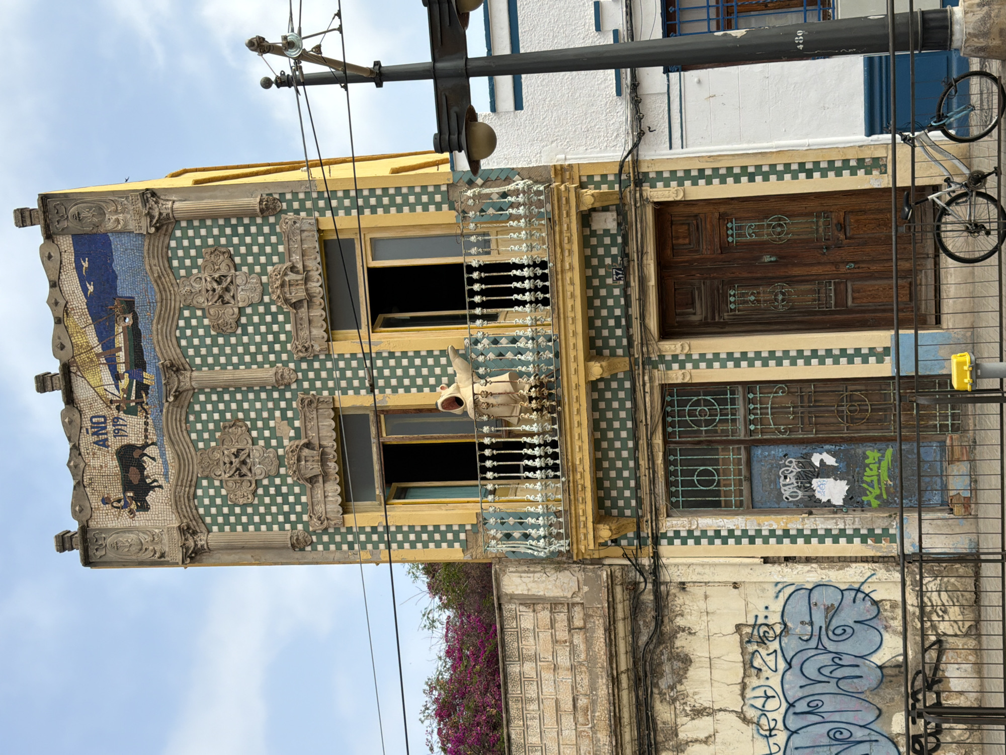 The image captures an art nouveau building facade on a sunny day in Unknown, Unknown. The building, adorned with green and white checkered tiles and yellow trim, features a mosaic depicting a bull and a ship at its apex, with the inscription "AÑO 1919". A balcony with ornate railings juts out from the second floor, where a figure in a light-colored robe stands with their arms outstretched. Below the balcony, two large wooden doors are visible, one of which is blue with graffiti. Adjacent to the building, another facade is covered in blue and white graffiti. A black metal fence runs along the front of the properties. A bicycle is parked near the fence, leaning against it. A lamppost with two lights stands on the right side of the frame. The sky is blue with scattered clouds.