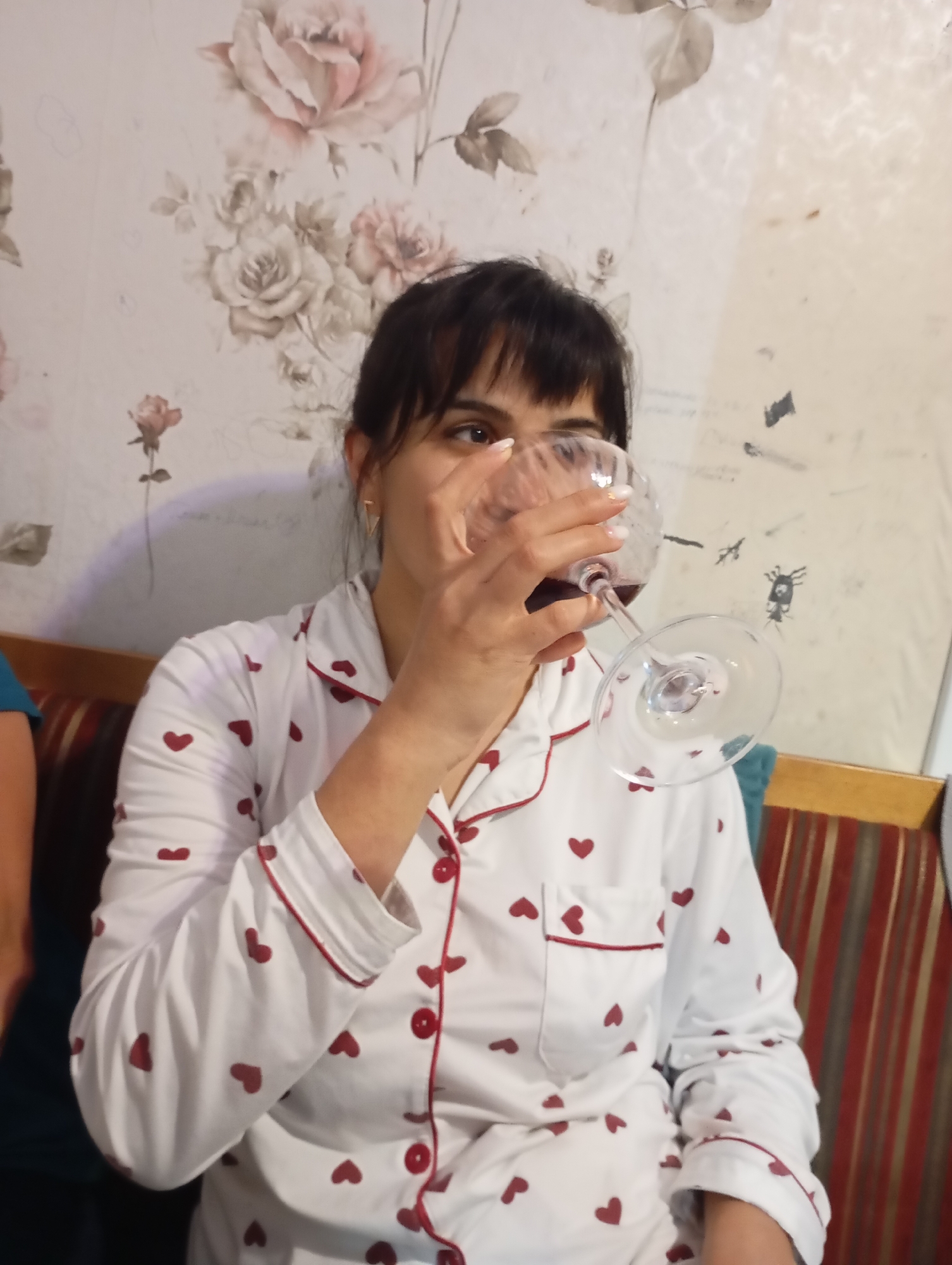 A woman with dark hair and bangs is seen from the chest up, holding a wine glass to her lips. She is wearing a white pajama top adorned with red hearts. The background features floral wallpaper with roses and leaves, along with a striped couch. The setting appears to be indoors.