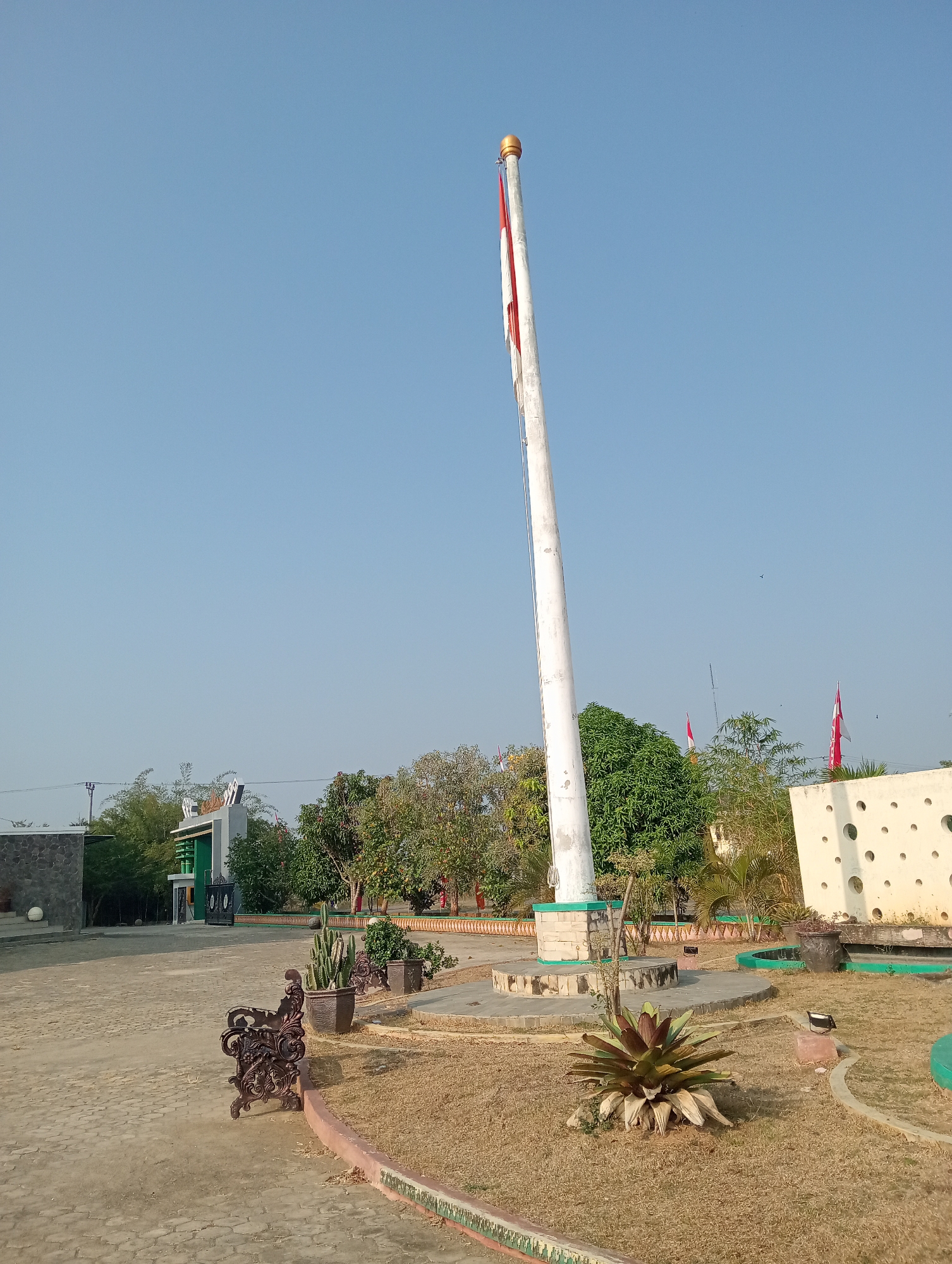 The image shows a large flagpole with an Indonesian flag flying in a park-like setting. The flagpole is tall and white, with a gold finial. The flag is red and white, the colors of the Indonesian flag. In the background, there is a green gate with ornate black gates, trees, and a white wall with circular cutouts. There is also a bench and several potted plants. The ground is paved with stones and dry grass. The sky is clear and blue. The location is likely the "Taman Wisata Alam Pringsewu" (Pringsewu Nature Tourism Park) in Pringsewu, Indonesia.