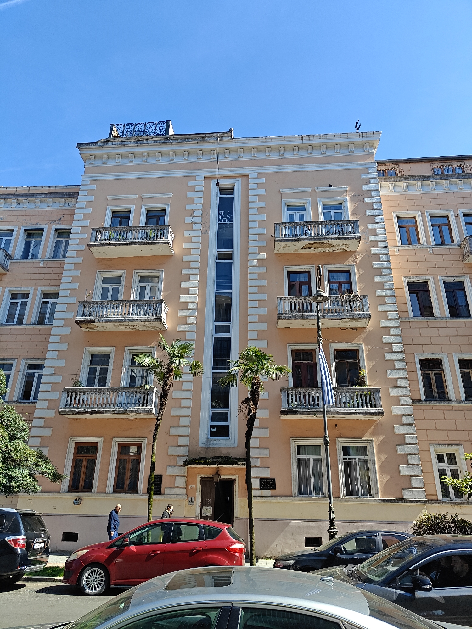 The image depicts a street scene in Batumi, Georgia. In the foreground, several cars are parked along the curb, including a red hatchback, a dark sedan, and a silver-grey sedan. Two pedestrians are walking on the sidewalk.
The focal point is a peach-colored building with white architectural details and balconies. A distinctive vertical window runs from the ground floor to the top of the building. A flag is visible in front of the building. The architecture style suggests a historical building. The sky is clear and blue.