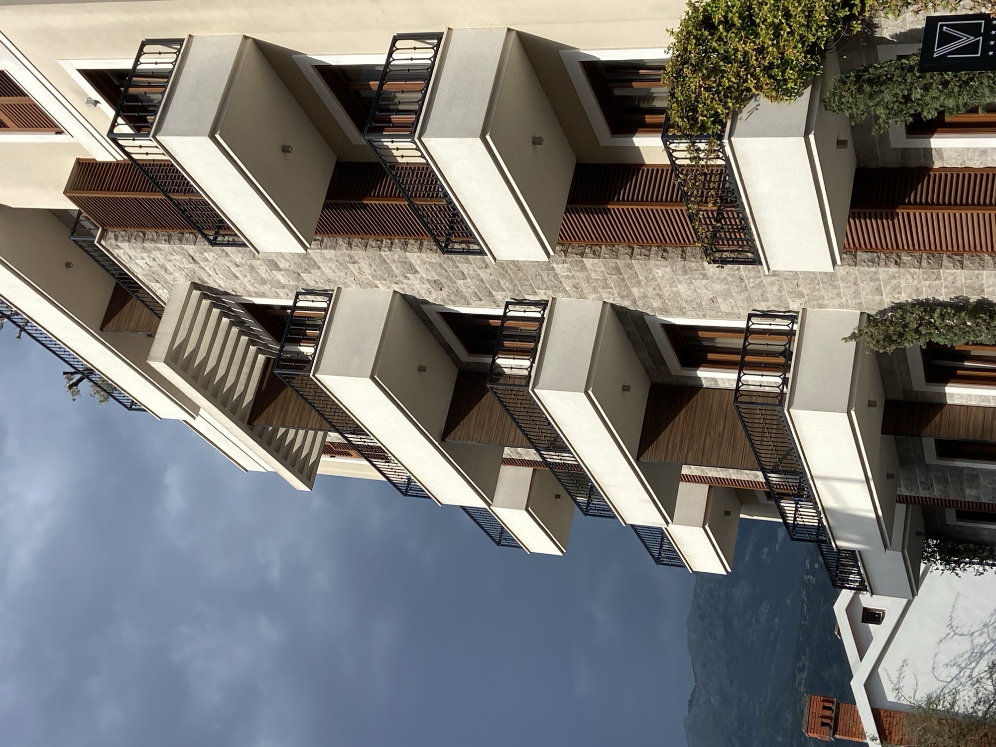 A building with a modern design is seen from below in Budva, Montenegro. The structure has protruding balconies with white walls and dark railings. Between the balconies are horizontal wood-paneled sections and areas of gray stone. Some balconies have greenery on them. The sky is overcast, and a mountain is visible in the distance. A neighboring building with a red brick chimney is partially visible.