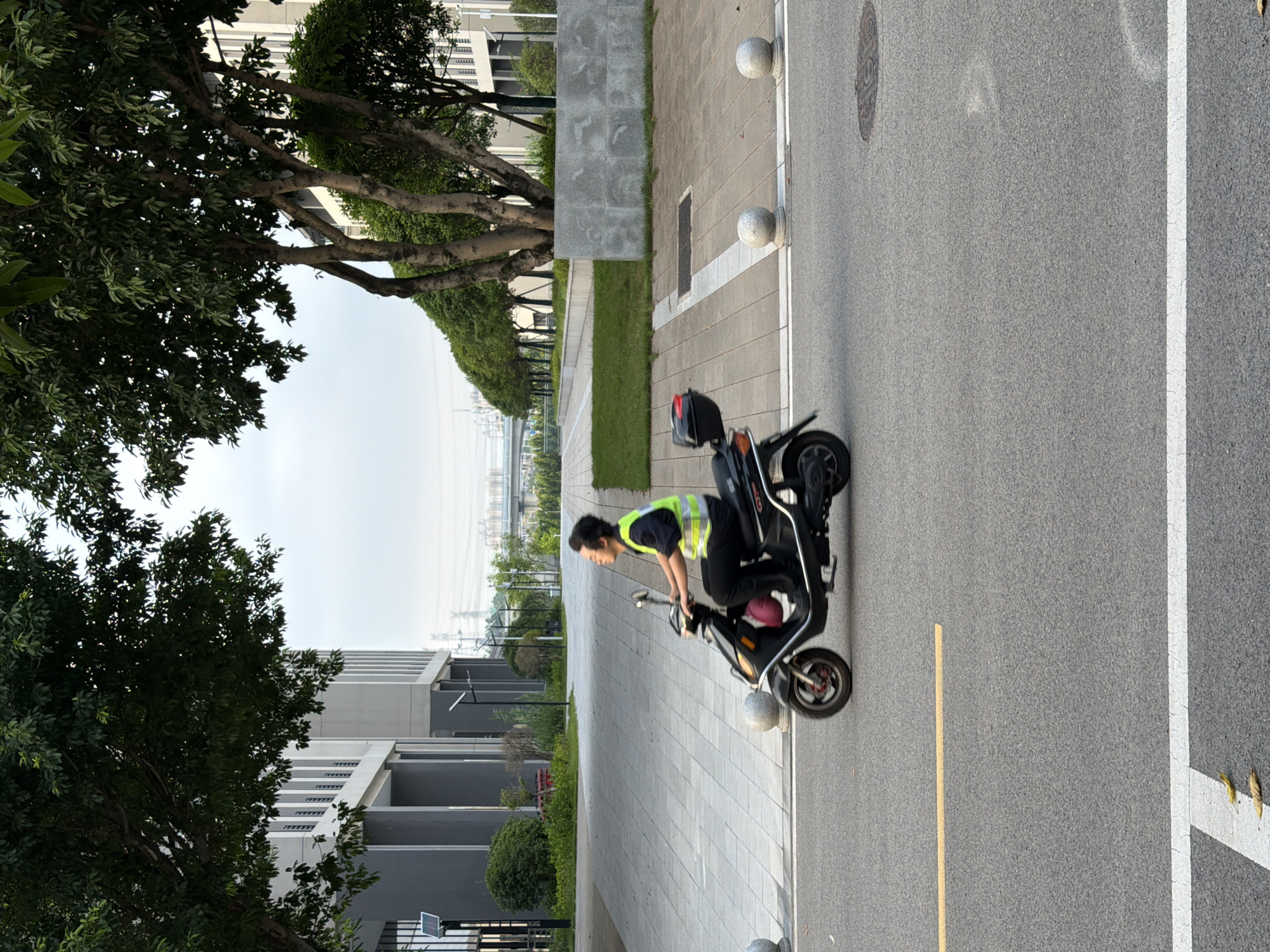 In a contemporary urban setting, likely within Jiangjin, China, a person wearing glasses and a high-visibility yellow vest over a dark shirt and pants rides a black electric scooter. The scooter has a red item secured to its rear. The rider is moving from left to right across the frame on an asphalt road, which features a single yellow line in the foreground and a white intersection marking. To the right of the road, a paved walkway is lined with spherical bollards, separating it from a grassy area and a modern building with large windows. Mature trees with dense green foliage are positioned in the middle ground, with more trees and vegetation lining a distant walkway. In the background, elevated structures and utility poles are visible against a hazy sky. The scene appears to be during daylight hours, possibly early morning or late afternoon, given the soft lighting.