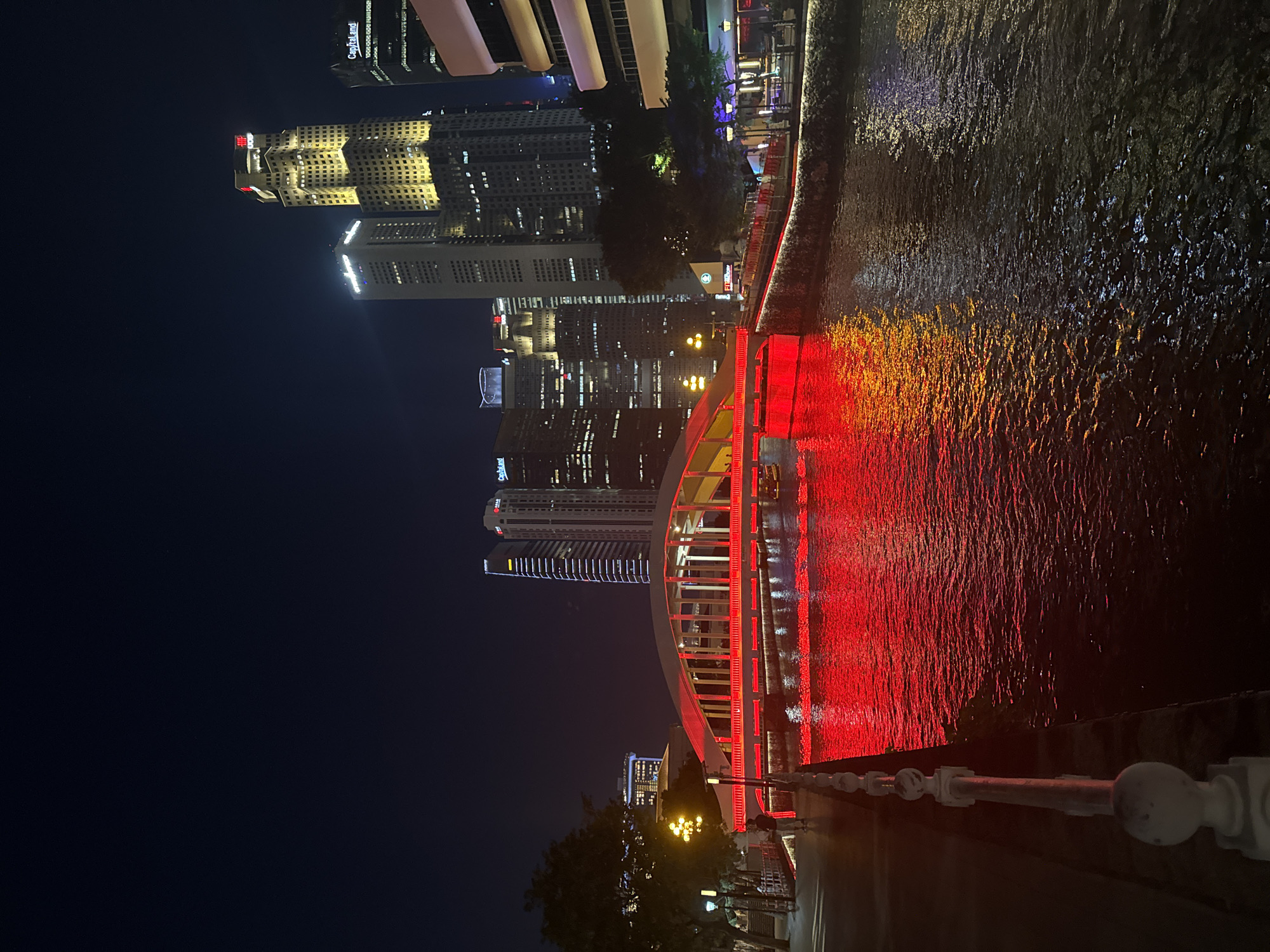 The image depicts the Singapore River at night, illuminated by bright red lights from a bridge spanning across the water. The bridge, a prominent architectural feature, has an arched design and is supported by pillars. The red lights reflect vibrantly on the dark water surface, creating a striking visual effect. Along the riverbank on the left, a walkway is visible with a white railing. A few streetlights and trees line the walkway.
In the background, a cluster of modern skyscrapers dominates the city skyline. These buildings are lit up, showcasing various architectural styles and illuminated windows. One tall skyscraper in the center features a golden glow, with prominent red signage at its peak. Another skyscraper to its right has the "Capitaland" logo visible. The sky is dark, characteristic of nighttime.
There are a few people visible on the walkway near the bridge, appearing to be strolling or observing the scenery. The overall scene conveys a sense of urban nightlife, with the illuminated bridge and buildings creating a lively atmosphere against the dark night sky and river.