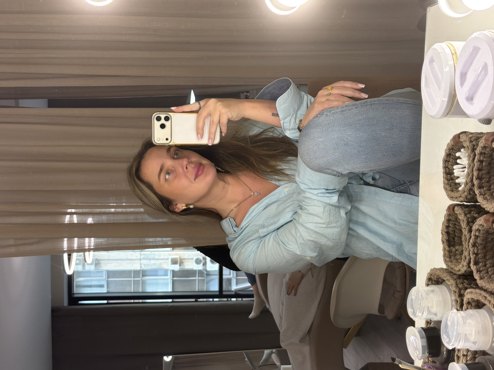A fair-skinned woman is taking a selfie in a room. She is holding a white smartphone. She has long blonde hair and blue eyes, with a necklace and earrings. She is wearing a light blue collared shirt and jeans.
The room has a window with a view of buildings, which suggests an urban location. In the mirror there are cosmetics containers and a decorative item. There are also makeup lights on the top of the mirror.
It is not possible to determine if the location is in Moscow, Russia from the provided image and details alone.