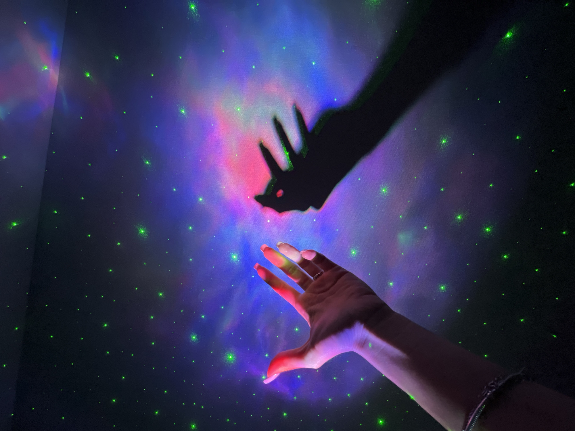 The image displays a hand with fingers outstretched, casting a shadow on a wall. The wall is illuminated with a galaxy projector, creating a starry, nebula-like effect with green, purple, and pink hues. The hand is lit from the side, casting a distinct shadow with four fingers visible. The overall scene evokes a sense of wonder and fantasy, as if the hand is reaching out to touch the cosmos. No specific location in Nizhny Novgorod, Russia, can be determined from the image.