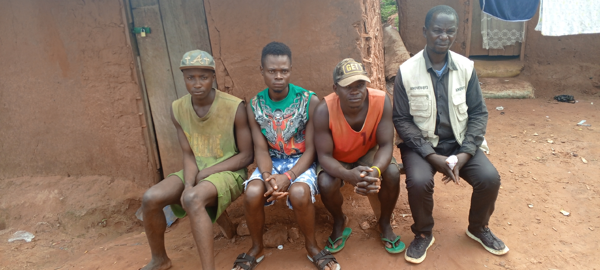Four men are sitting together outside a structure with a wall made of earth or clay bricks. The man on the far right wears a white vest over a dark long-sleeved shirt and dark trousers, and black sneakers. The man next to him wears an orange tank top and brown shorts. He has a backwards hat. The man next to him is wearing a green t-shirt and blue denim shorts. The man on the left is wearing a green t-shirt and green shorts and hat. There is a door visible in the background to the left, secured with a padlock. Another structure can be seen behind the man on the right, with laundry hanging. The ground is dirt. The location is in Makungu, Democratic Republic of the Congo.
