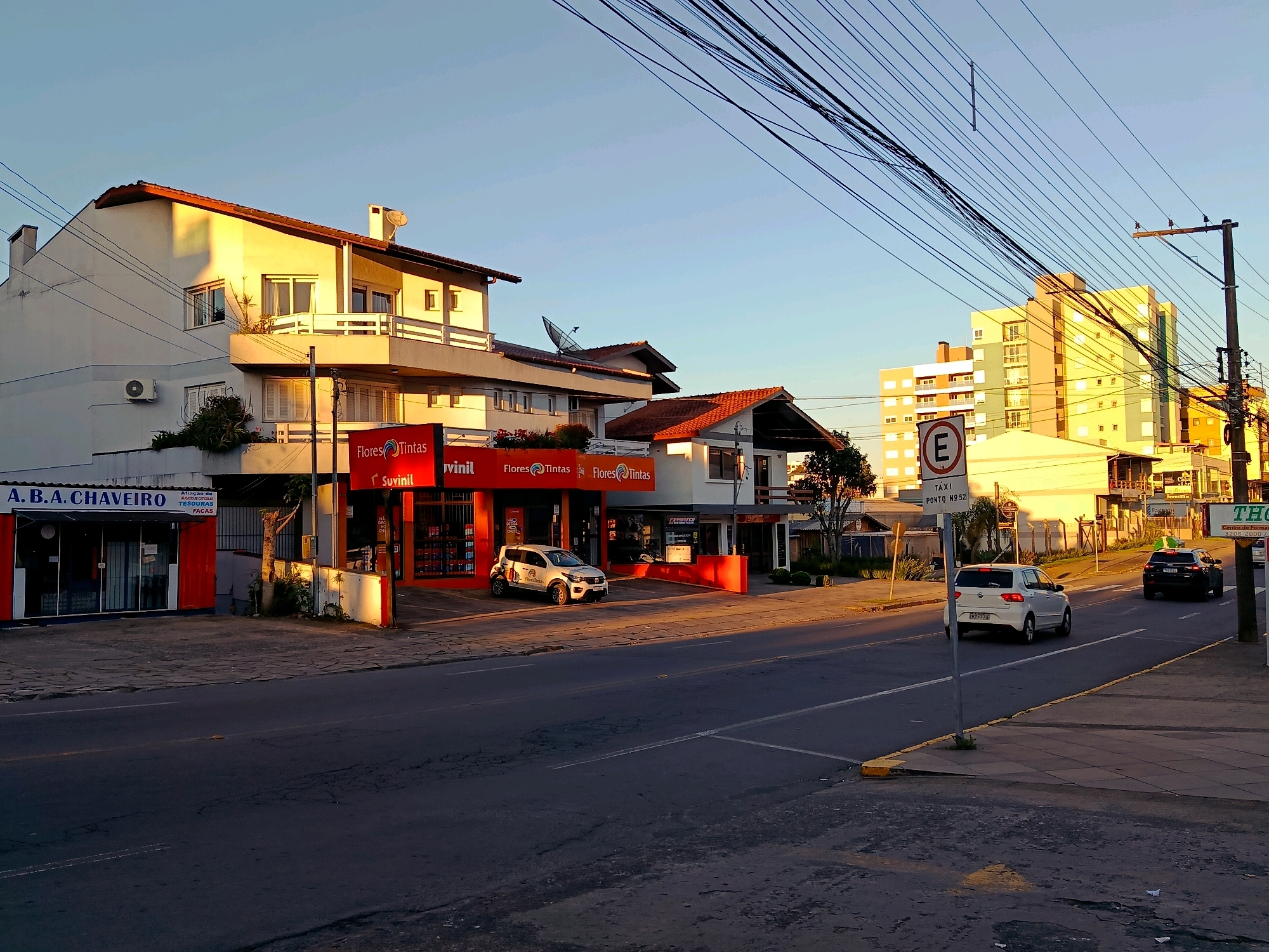 The image depicts a street scene in Caxias do Sul, Brazil, likely during the late afternoon or early evening, as evidenced by the long shadows and warm sunlight illuminating the buildings. The perspective is from the side of the road, looking towards a row of businesses and multi-story buildings.
On the left, a store with the name "A. B. A. CHAVEIRO" is visible, featuring a red facade and large glass windows. Adjacent to it, there is a store with a bright red exterior, branded with "Flores Tintas" and "Suvinil," suggesting it sells paints and related products. A white hatchback car with "Flores Tintas" branding is parked in front of this store.
Further down the street, the road curves slightly. Several multi-story buildings are visible, some with modern architectural styles featuring large windows. One prominent building in the background is a colorful, tall structure with yellow and green sections.
Traffic signs are present, including a white circular sign with a red border and the letter "E" inside, indicating parking restrictions. Another sign reads "TÁXI PONTO Nº 52," marking a taxi stand. Cars are visible on the road, including a white hatchback and a black SUV driving away from the viewer. Electrical wires are strung across the sky, supported by utility poles, a common sight in urban environments. The sidewalks are paved, and some vegetation is present, including small trees and bushes. The overall impression is of a commercial and residential street in an urban setting.