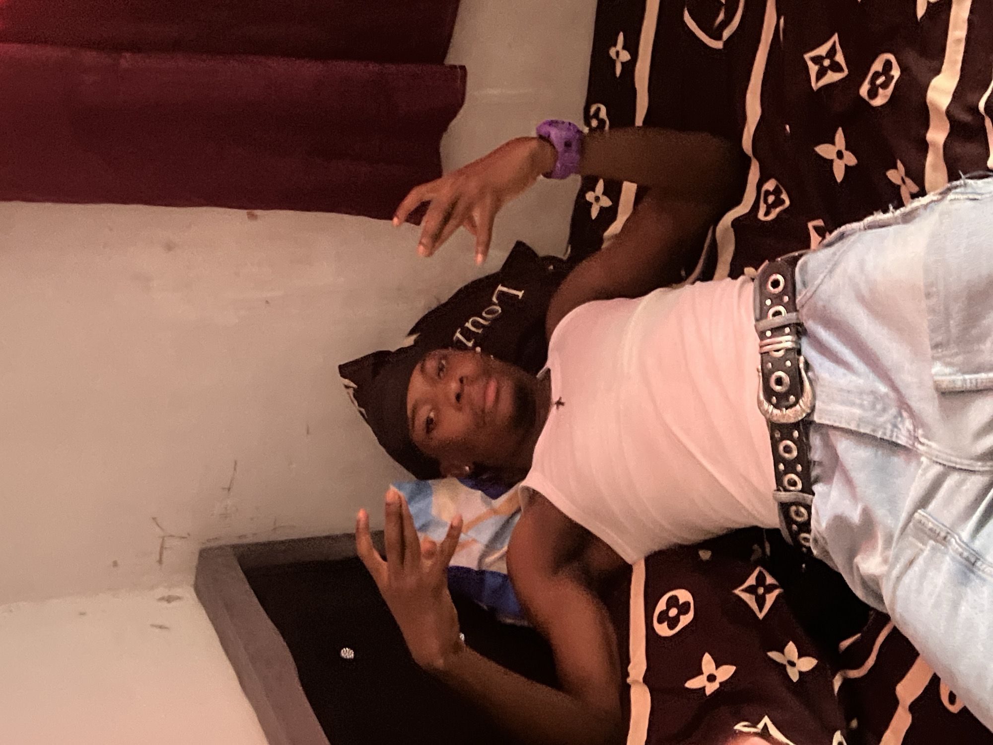 A young Black male is lying on a bed, propped up against a headboard. He is wearing a white tank top, light-wash jeans, a black bandana, and a black belt with a silver buckle. He has a purple watch on his left wrist. His right hand is raised, palm facing forward, with his index and pinky fingers extended, and his middle and ring fingers curled. His left hand is in a similar pose. The bed has a patterned comforter with a floral design and what appears to be the Louis Vuitton logo. The background includes a white wall with some discoloration and peeling paint, and a dark red curtain. The scene is indoors, likely a bedroom. The location is not identifiable as Durimi, Nigeria, based solely on the visual information provided.