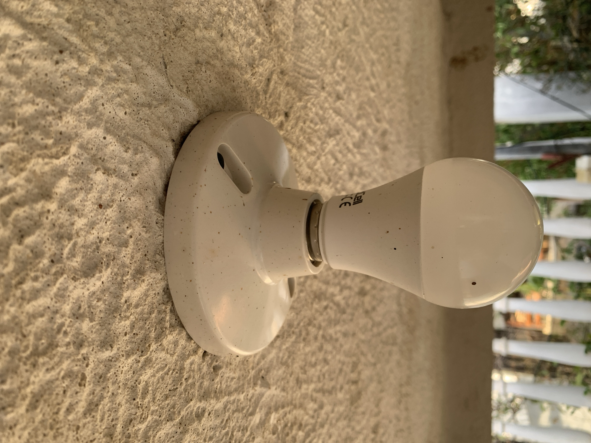 An illuminated light bulb is affixed to a roughly textured wall of a pale, neutral color. The light fixture is a simple, circular white mount with holes for screws. The bulb is white and has visible markings and slight surface imperfections. Beyond the bulb and wall, blurred greenery and a white shuttered window or fence are visible, suggesting an outdoor setting.
I cannot provide the location in the city of Santa Ana, Venezuela based on the visual information provided.