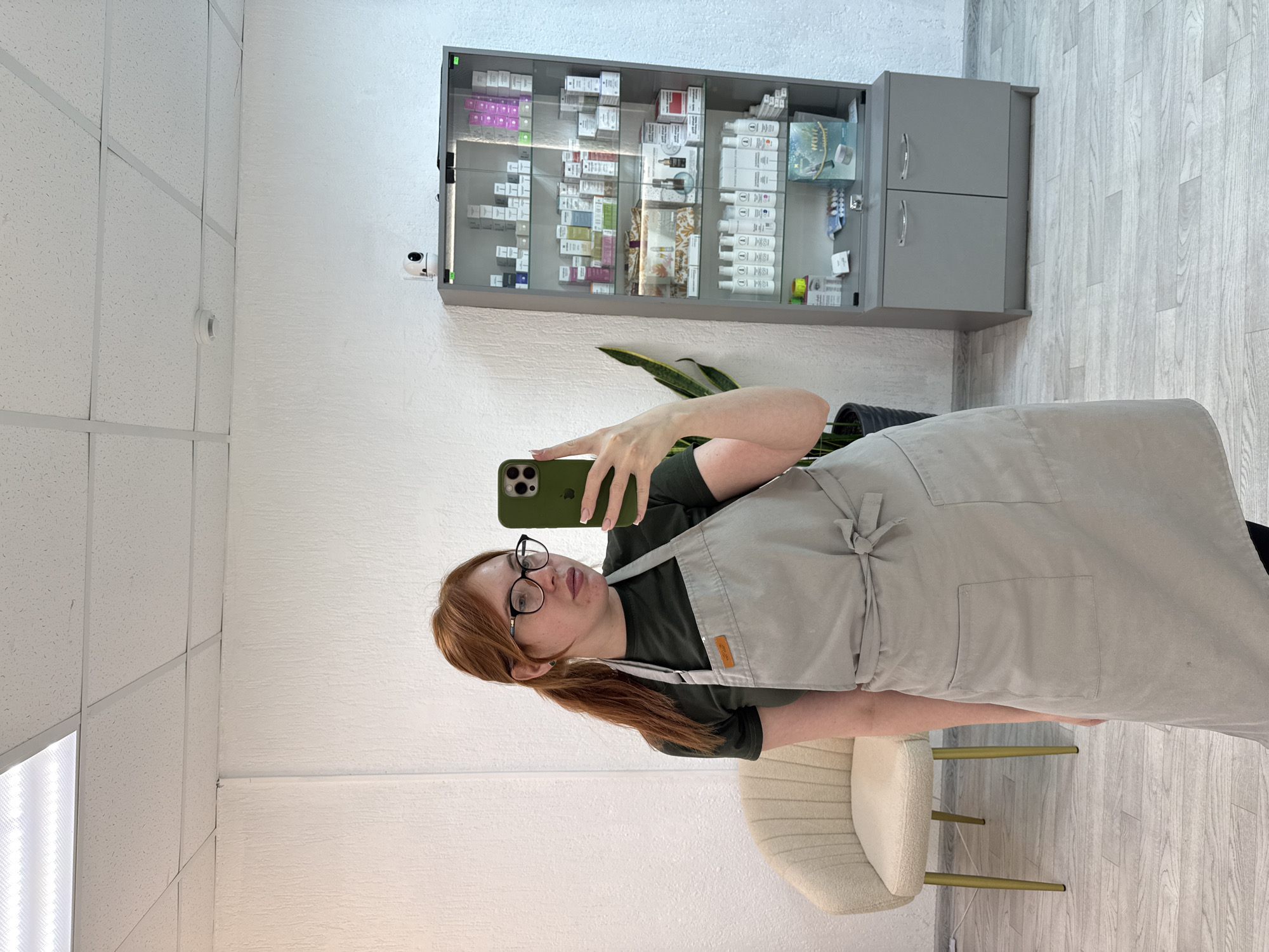 An indoor selfie captures a young woman with reddish-brown hair, tied in a ponytail, wearing black-rimmed glasses, a dark green t-shirt, and a light gray apron. She is holding a green smartphone, seemingly an iPhone, to take the picture, reflecting her neutral expression.
The setting appears to be a professional or commercial space, possibly a beauty salon, clinic, or shop in Chaikovsky, Russia. The background features white walls with a textured finish and a white acoustic tile ceiling. The floor is covered with light-colored wood-effect laminate or vinyl.
Prominently displayed on the wall behind the woman is a gray cabinet with glass doors, filled with various cosmetic or medical products. These include numerous small boxes in different colors (pink, green, blue, white), as well as white bottles, some of which appear to have "NANO" visible on their labels, and a blue box with a "WOM" or "WOK" logo. A small white security camera is mounted to the wall just left of the cabinet. Below the cabinet, a potted plant with large green leaves is visible. To the right, a cream-colored upholstered chair with golden legs sits on the wood-effect floor. A white smoke detector is visible on the ceiling.
The activity is a selfie being taken in what appears to be a workplace. The interior is brightly lit, suggesting daytime. The woman's calm demeanor and the organized professional environment contribute to a mundane, everyday scene.