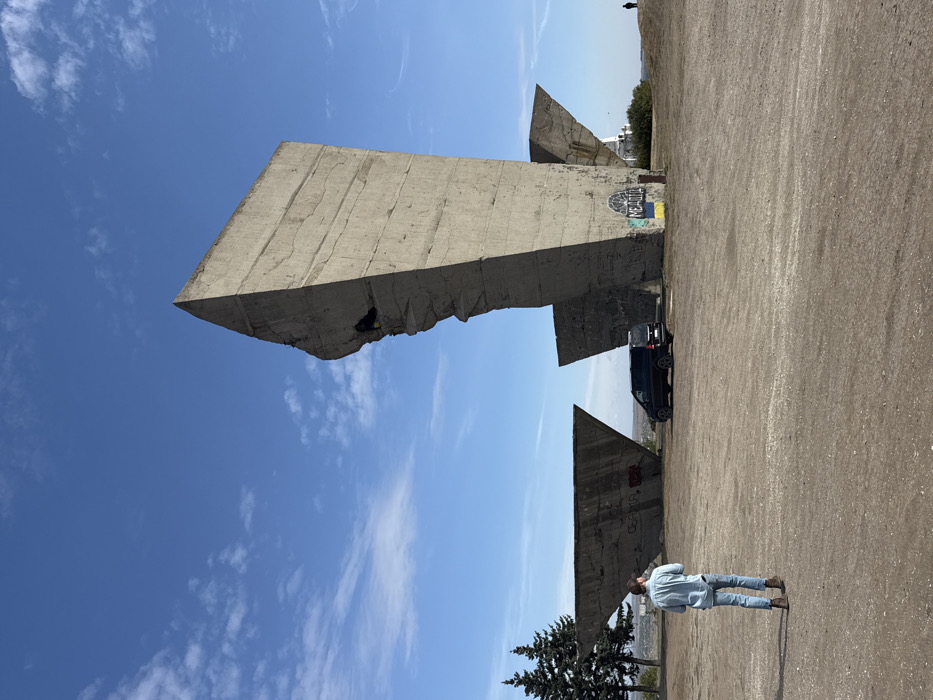 The image displays the "Monument to Liberators" located on Mount Kremenets in Izium, Kharkiv Oblast, Ukraine, under a clear blue sky with scattered white clouds. The primary subject is a large, weathered concrete monument composed of multiple angular, Brutalist-style slabs situated on unpaved light brown earth. The most prominent slab, centrally positioned on the right, is tall and angular, featuring visible horizontal striations from its construction and irregular, jagged damage along its left vertical edge. The base of this slab displays spray-painted graffiti: the word "МЕДОВА" (MEDOVA) above a blue and yellow Ukrainian flag. To the left, a lower, triangular concrete slab also bears faint graffiti, including "СЛАВА" (SLAVA) and "ВОЛЯ" (VOLYA). A dark-colored Mercedes-Benz van is parked facing left on the dirt ground between these two concrete elements. In the foreground, a male individual is captured from behind, walking away from the viewer towards the monument. He wears a light blue long-sleeved shirt, blue jeans, and brown boots. Two evergreen trees are visible on the far left. A faint, distant figure is discernible on the far right horizon, and the background shows a flat horizon line over what appears to be a distant landscape or city.