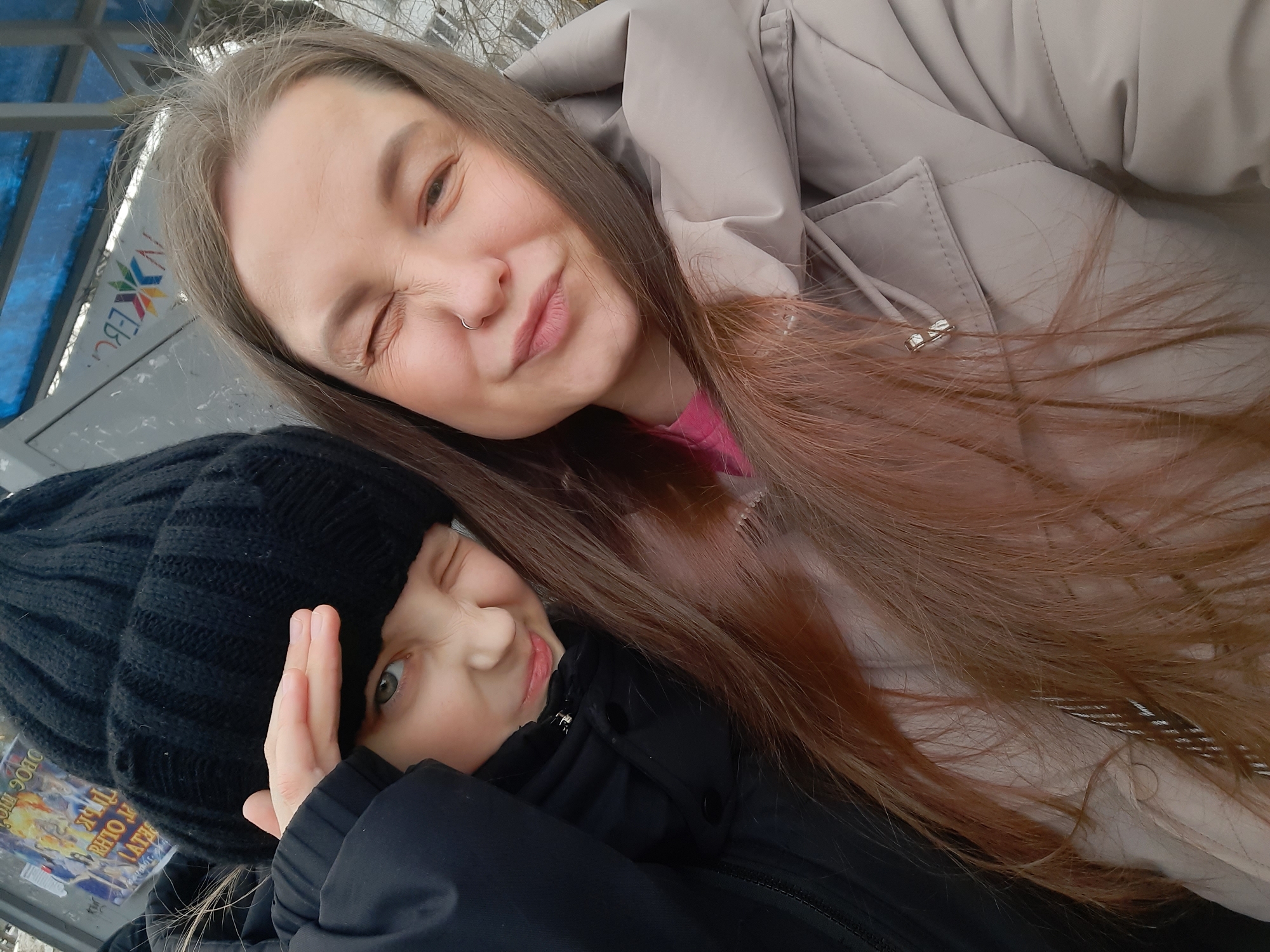 Two individuals, likely a woman and a child, are posing for a selfie-style photograph outdoors. The woman is positioned at the top of the frame, wearing a beige puffer jacket and a pink shirt underneath. She has long, light brown hair, fair skin, and is winking with her right eye. She has a piercing on her left nostril. The child is below her, wearing a black winter hat and a black jacket. The child is also winking with their right eye and has their left hand raised near their forehead. The background includes elements of a city bus stop or public transportation shelter, identified by the presence of a blue roof and advertisement with colorful lettering: "NXEBC." The location is Izhevsk, Russia.