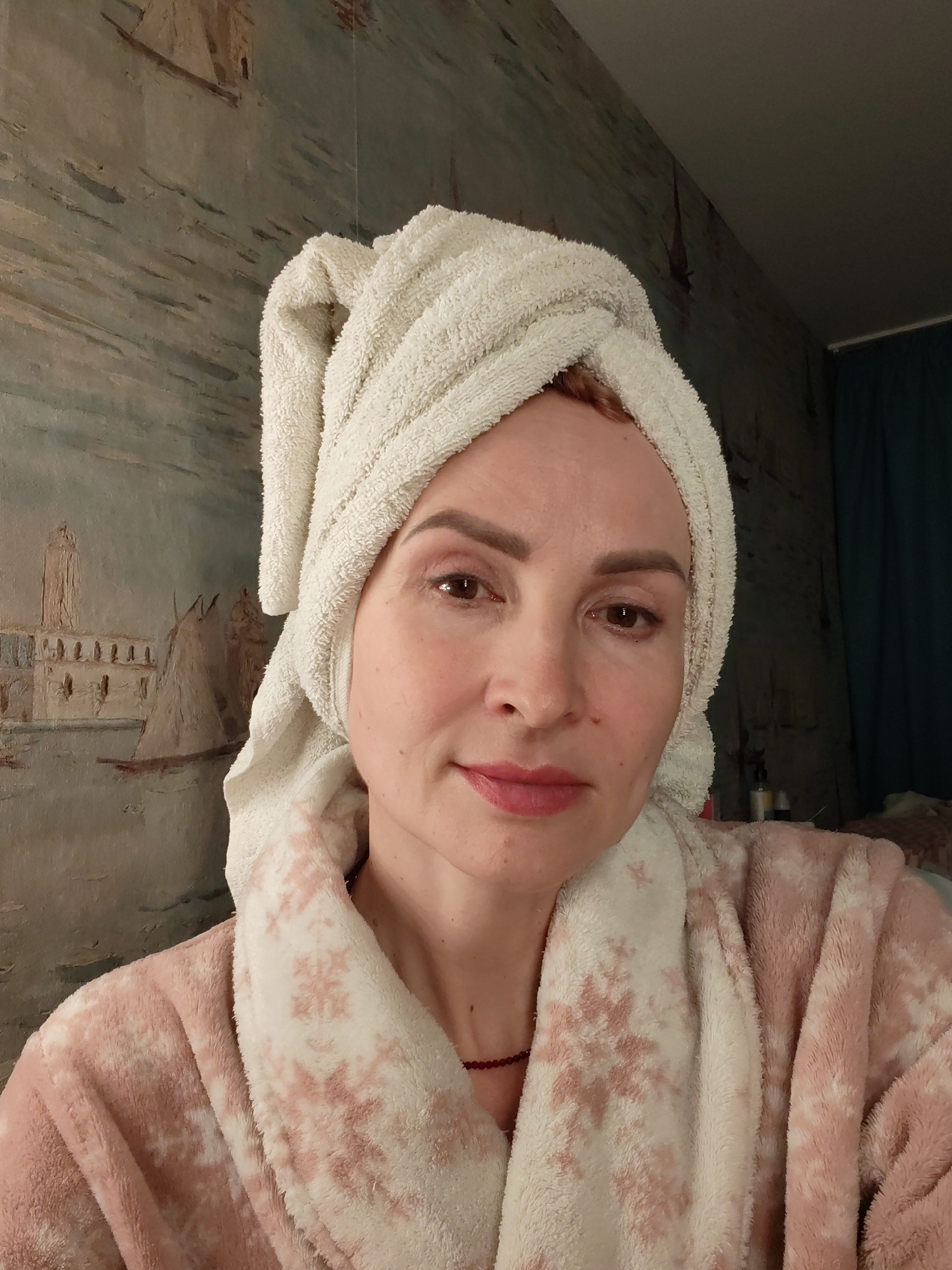 A woman is shown in a close-up selfie. She is wearing a pink and white patterned robe and a white towel is wrapped around her head. In the background is a wall decorated with a mural depicting a waterscape with buildings. Based on visual information alone, I cannot determine if this image was taken in Chaikovsky, Russia. There is no identifying information in the image that allows that specific location to be verified.