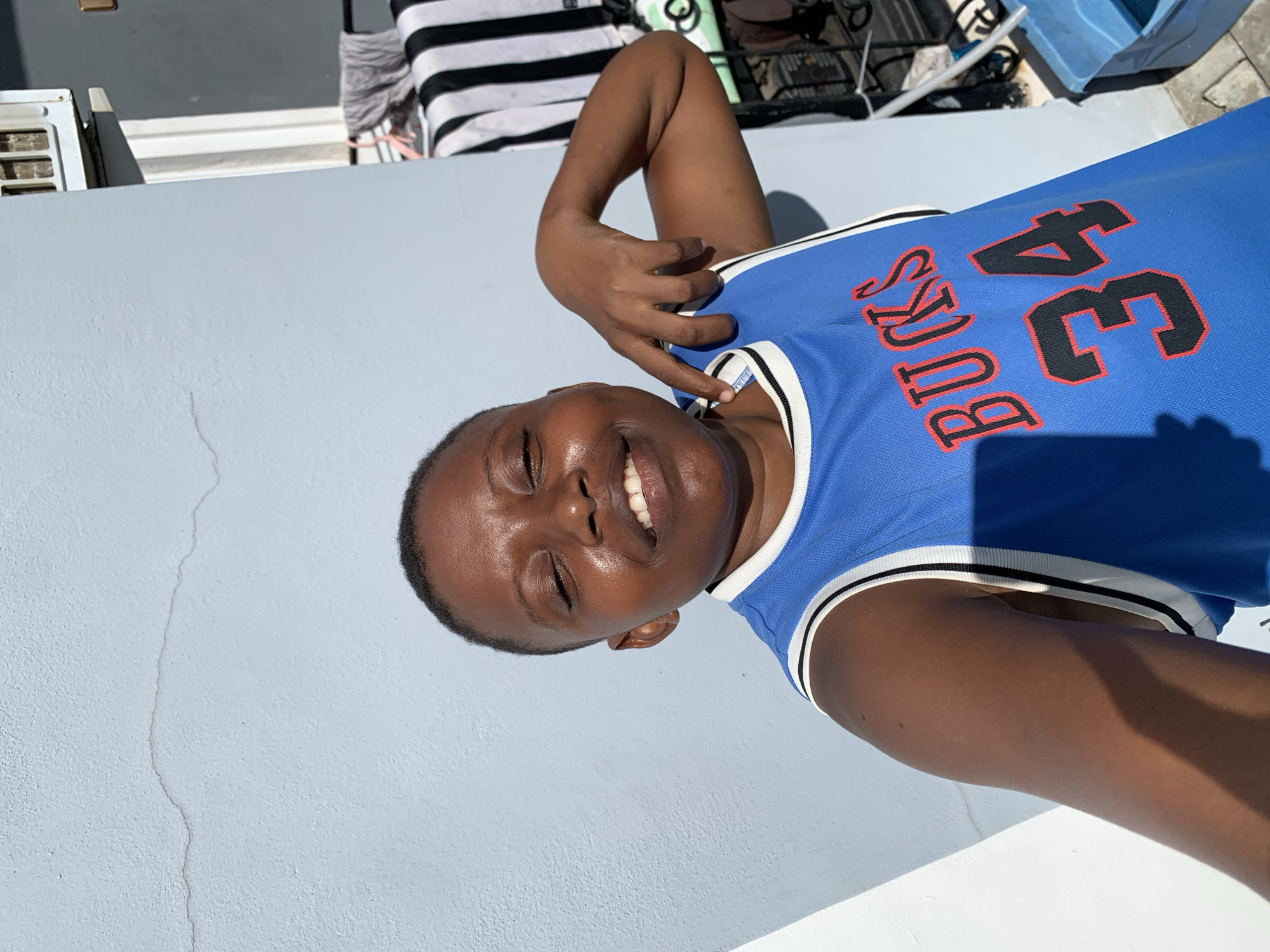 A young person with dark skin and short dark hair is captured in a casual outdoor portrait, rotated counter-clockwise in the image. They are wearing a bright blue basketball jersey featuring "BUCKS 34" printed in red with a black outline across the chest, and white and black striped trim on the collar and armholes. The person is smiling broadly with their eyes closed, conveying happiness and contentment, possibly squinting from the bright sun. One hand is raised, with fingers lightly touching their neck or ear.
The background features a light blue-gray wall, which displays a thin vertical crack in the lower left portion. In the upper background, outdoor elements are visible, including part of an air conditioning unit or vent, a piece of black and white striped fabric (possibly a towel or clothing), and a blue plastic container. The strong, direct sunlight casts distinct shadows, indicating a clear, sunny daytime. The scene appears to be an outdoor area, such as a patio, balcony, or rooftop, located in Ibasa, Nigeria.