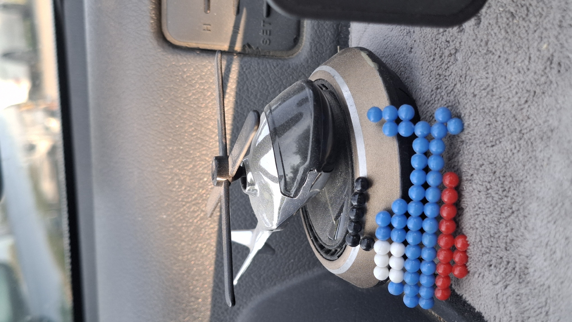 A close-up, eye-level, medium shot shows a toy helicopter with a rotor and tail rotor, mounted on a circular base with what appears to be a solar panel. The helicopter is silver and black. In front of the helicopter, a small mosaic helicopter shape is made of colorful plastic beads: white, blue, and red. The mosaic helicopter is arranged in horizontal rows of beads, with the tail and a portion of the body made of blue beads, the body section above that made of white beads, and the front section and landing gear made of red beads. The helicopter and bead mosaic are positioned on a textured gray surface, possibly a car's dashboard or floor mat, within a vehicle. The background is blurred and shows a dashboard and possibly a windshield reflecting an outdoor scene. The image does not contain any information about the city of Shymkent, Kazakhstan.