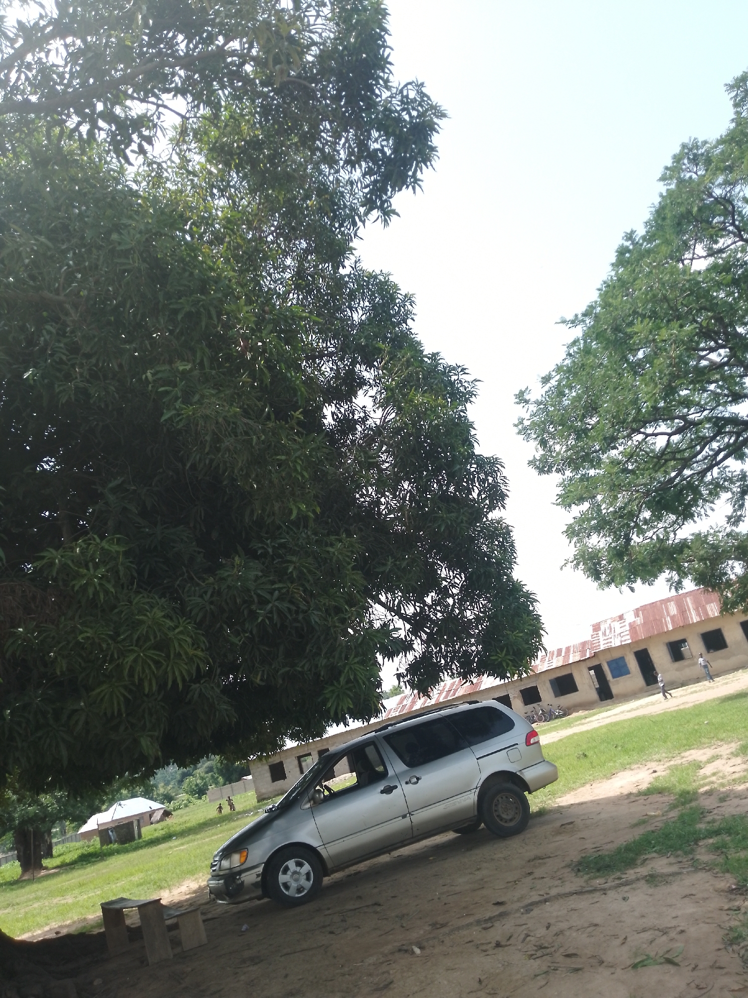 In Serti, Nigeria, a silver minivan is parked on a dirt path under a large, leafy tree. The minivan is angled towards the viewer's left, with its front right tire slightly off the ground due to the uneven terrain. To the right of the minivan, a long, single-story building with a corrugated metal roof and numerous windows stands in the background. Several figures, appearing to be children, are visible in front of this building. The environment is rural, with green grass and more trees visible.
