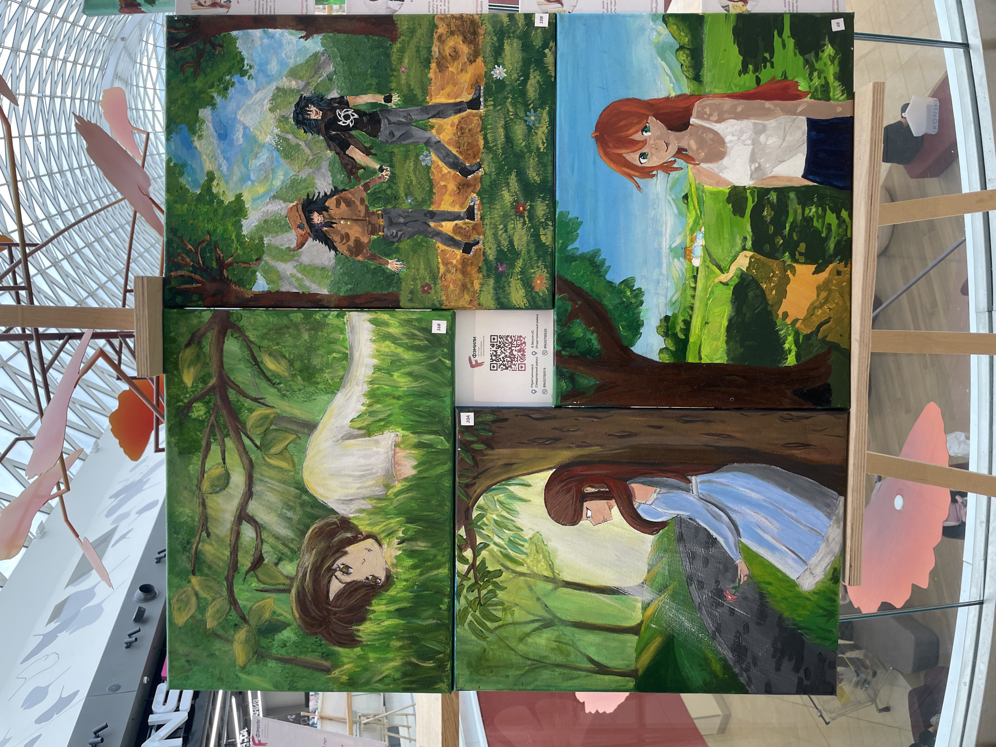 An indoor display of four anime-style paintings is shown, set up on a wooden easel. The paintings are arranged in a 2x2 grid. The top left painting depicts a person with short brown hair lying in green grass, with their head resting on their arm. Their eyes are wide open and looking towards the viewer. The top right painting shows two figures with dark hair walking hand-in-hand on a path through a grassy area with trees and a mountain landscape in the background. The figure on the left wears a brown jacket and hat, while the figure on the right wears a black t-shirt. The bottom left painting features a person with long brown hair wearing a light blue dress, standing in a sunlit forest with a pink flower in their hand. The bottom right painting portrays a person with long orange hair wearing a white tank top and a dark blue skirt, standing in a green meadow with houses in the distance. The background of the scene appears to be a shopping mall with a glass ceiling and some pink decorative bird sculptures hanging overhead. A sign in Russian is visible to the left of the display, which is located in the city of Perm, Russia.