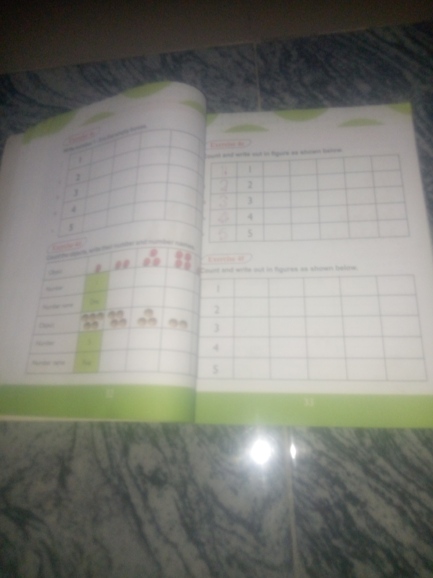 The image shows an open book with exercises related to numbers. The left page has a table with rows labeled 1 through 5 and columns. Below this table, there is another section with rows labeled "Object", "Number", and "Number name". This section contains images of dots arranged in groups, and corresponding numbers and number names are written. For example, there are images of 1, 2, 3, and 4 dots, and the number names "One" and "Two" are visible. The right page shows a similar table with rows labeled 1 through 5, but this table appears to be empty, intended for the user to fill in. The book is placed on a surface that has a patterned texture, possibly a table or floor. The lighting in the image is not uniform, with a bright glare visible in the lower right portion. There is no indication of the city of Jauro Yinu, Nigeria, or any specific location.