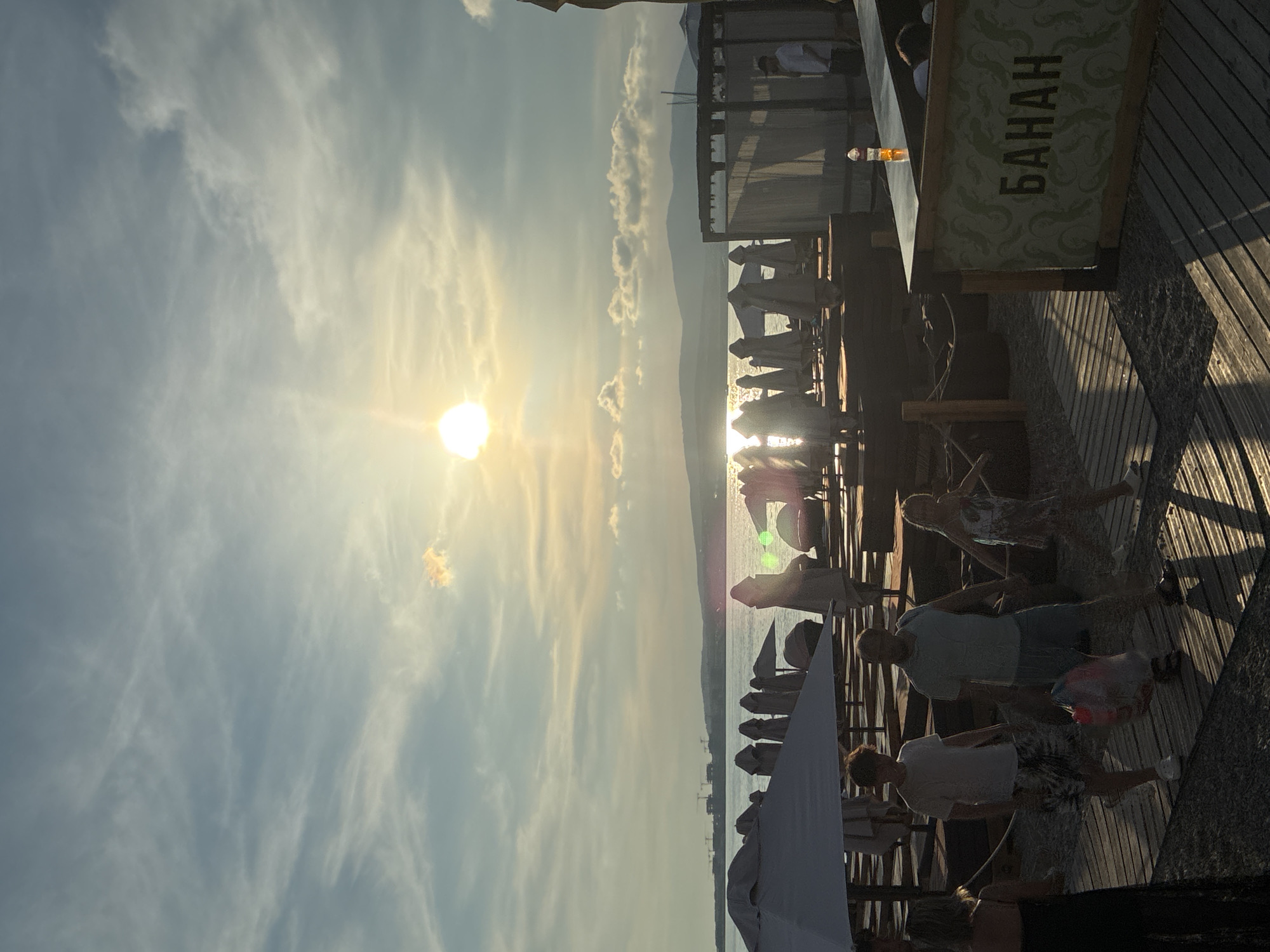 The image depicts a beachside scene at sunset in Gelendzhik, Russia. The sun is low in the sky, casting a warm glow and creating lens flare. Several people are visible walking on a wooden boardwalk near the water. In the foreground, a sign with the word "БАНАН" (Banana) is visible. Umbrellas are set up on the beach, suggesting a recreational area. The sea is calm, and hills are visible in the distance. Construction cranes are also present on the horizon, indicating ongoing development. The overall atmosphere is one of a relaxed evening by the sea.