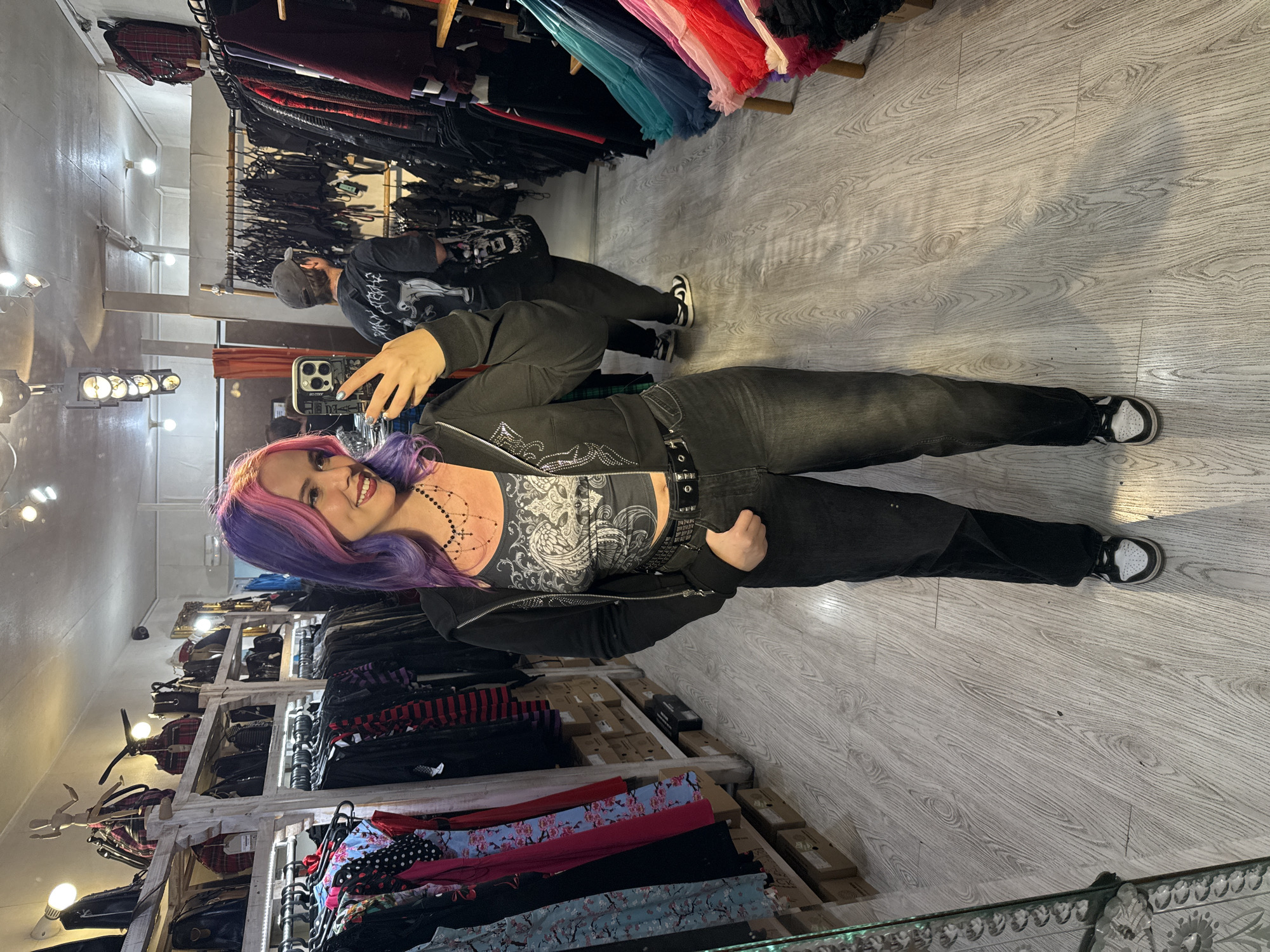 The image is a selfie taken by a woman with pink and purple hair in what appears to be a clothing store. She is wearing a patterned tank top, a black zippered jacket, black jeans, and black and white sneakers. The woman is smiling and holding a smartphone up to take the picture. In the background, another individual is seen looking at clothing racks. The store is well-lit with wooden floors and racks filled with various items of clothing. There is no readily identifiable information to indicate that the location is Madrid, Spain.