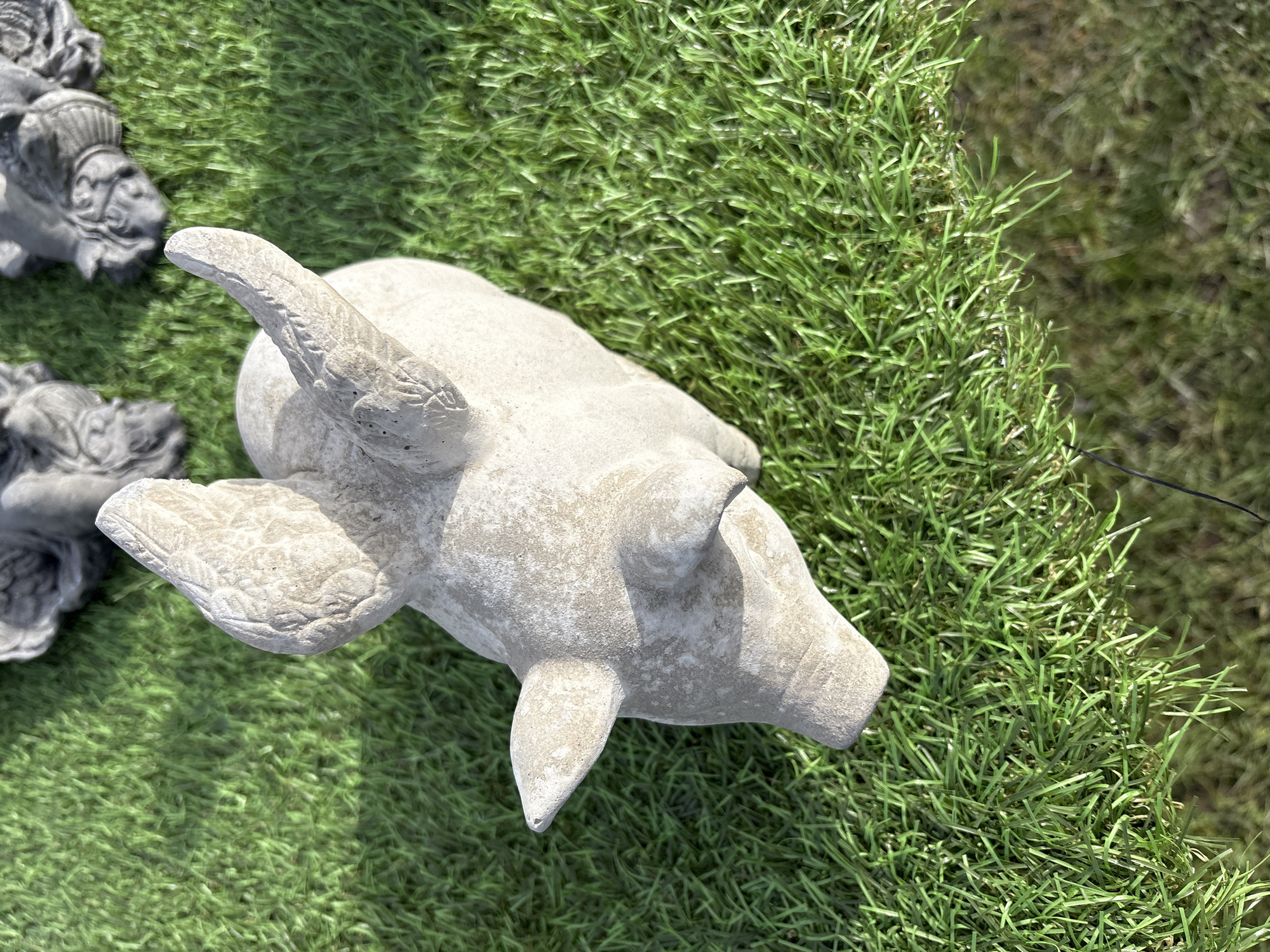 An aerial view shows a cement garden statue of a pig with wings on a patch of bright green artificial turf in Chirk, United Kingdom. Another, darker garden statue is partially visible at the left side.