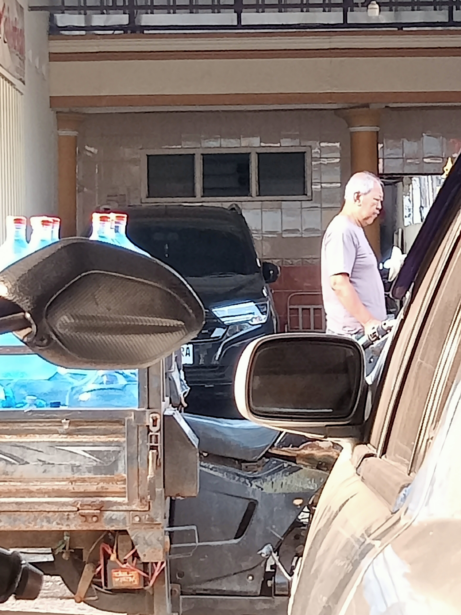 The image shows a man with grey hair and a light purple t-shirt standing near the open driver's side door of a light-colored car. He is holding a black power tool and appears to be working on the car. In the background, there is a dark SUV and several large blue bottles. A motorcycle mirror is visible in the foreground. The scene appears to be in an outdoor area, possibly a driveway or a shop, with a building in the background. The lighting suggests it is daytime. The location is identified as Simo, Indonesia.