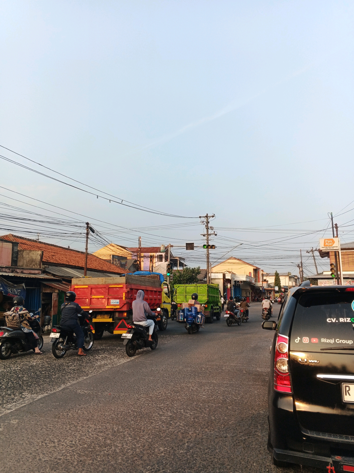 The image captures a street scene in Cilacap, Indonesia, during what appears to be late afternoon or early evening, given the warm light. The perspective is from inside a vehicle, looking forward. The road is paved and shows signs of wear.
In the foreground, a black SUV is partially visible on the right side of the frame. Its rear taillight assembly is prominent. A license plate reading "R" is visible. Decals on the SUV include social media icons for Instagram and YouTube, along with the text "CV. RIZQ GROUP".
The street is moderately busy with traffic. Several motorcycles are present. On the left, a black scooter with a rider wearing a patterned jacket and dark pants is parked. Further ahead, two motorcycles are in motion, each with a single rider. One rider is wearing a blue jacket and dark pants, while the other wears a grey hooded sweatshirt and white pants.
Dominating the mid-ground are two trucks. The first, a red dump truck, is stationary or moving slowly. Its license plate is visible, reading "0049B". The second truck, a bright green dump truck, is positioned behind the red one and is also in motion or stopped.
Along the sides of the street, buildings are visible. These appear to be shops or residential structures with tiled roofs. Various signs are affixed to the buildings, including one with "Pos" and "KURIPAN" visible.
The sky is a pale blue with some wispy clouds, suggesting fair weather. A complex network of overhead power lines stretches across the scene, supported by utility poles. Traffic lights are visible, with one showing a green light. The overall impression is a typical urban street environment in Indonesia.