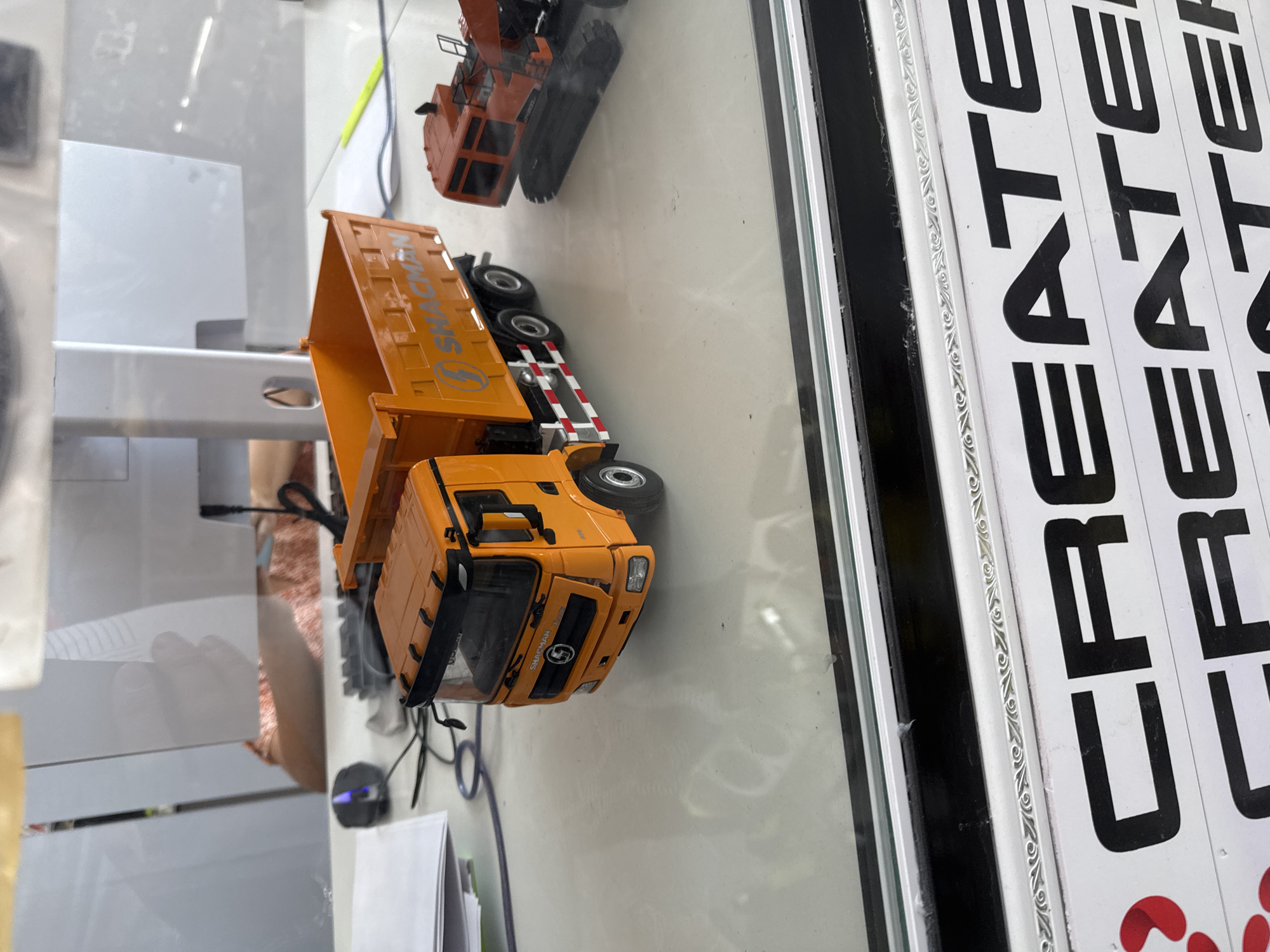 A toy dump truck and a toy excavator are displayed on a white surface, likely a desk, with a glass barrier in the foreground. The dump truck is orange and black, with "SHACMAN" written on its side. The excavator is orange and black with a track base. In the background, a computer monitor and a hand are visible, suggesting an office or retail environment. There is a partial view of text at the bottom of the image, appearing to be part of a sign or display. The location is identified as Shymkent, Kazakhstan.