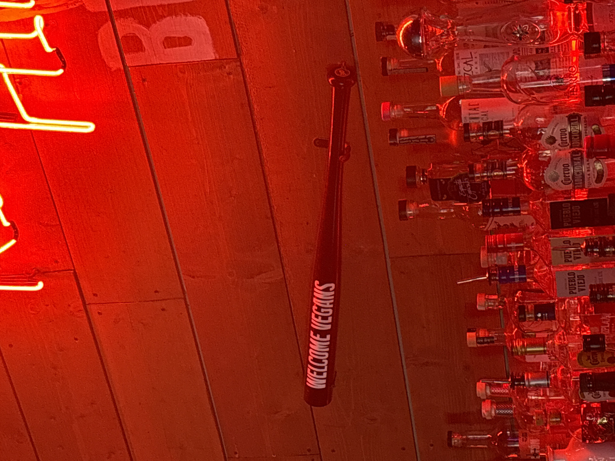 The image shows a partially lit wall with a neon light, a "Welcome Vegans" baseball bat, and a wall of liquor bottles. The wood paneling is visible behind the neon light and baseball bat. The baseball bat is dark and has the words "Welcome Vegans" printed in white on the side. The liquor bottles are arranged in rows and include various brands and types of alcohol, like Cuervo, Mezcal and Pueblo Viejo. The photo appears to be taken in a bar or restaurant.