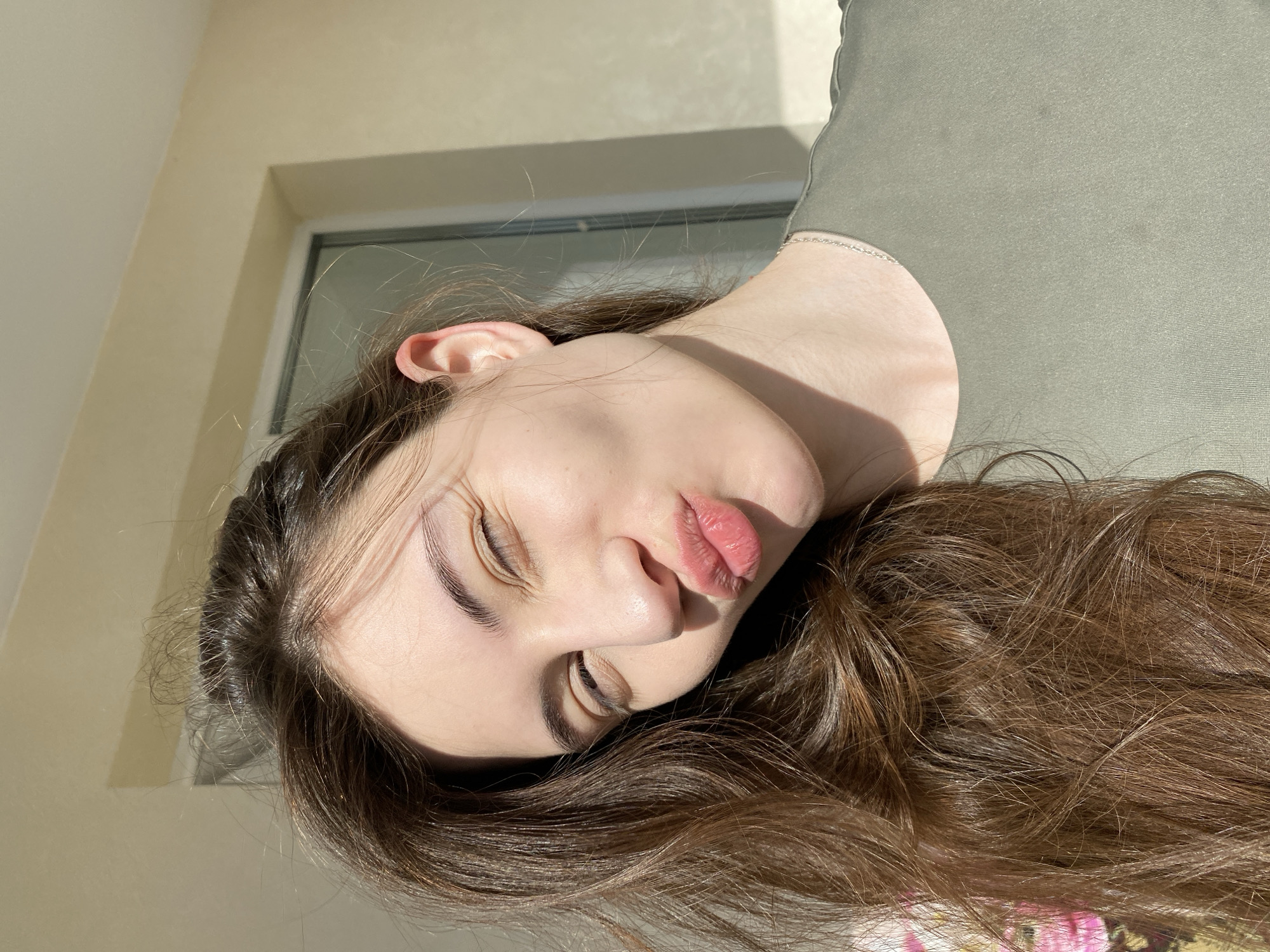 An eye-level portrait, rotated 90 degrees counter-clockwise, features a young woman with her eyes closed and lips puckered in a playful or affectionate expression. Her long, wavy brown hair cascades around her head, framing her face and extending downwards across the bottom of the frame. She has well-defined eyebrows and appears to be wearing light makeup.
The woman is illuminated by strong, direct sunlight, which creates prominent shadows across her cheek, neck, and the light-colored wall behind her. This suggests the photo was taken during the daytime, possibly morning or late afternoon, on a bright, sunny day. She is wearing an olive green garment, likely a top or dress, visible on her upper body.
In the blurred background, part of a window frame with a grey or white edge is visible, set against a light, possibly cream or beige wall. The scene is indoors, in what appears to be a room. There is no visible text in the image. The image captures a moment of playful posing, likely in Votkinsk, Russia.