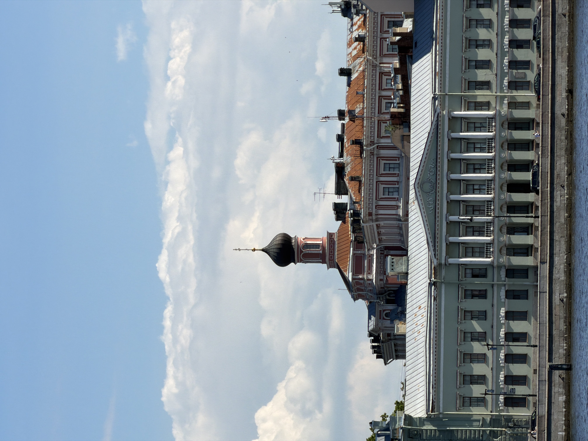 The image, when rotated upright, displays a sunny daytime scene of architectural landmarks along a waterway in Saint Petersburg, Russia. The sky is bright blue with scattered, fluffy white clouds, suggesting fair weather.
On the right side of the frame, a calm body of water, likely a river or canal, stretches horizontally, bordered by a sturdy stone embankment. Several dark cars are parked along the embankment, and a few classical street lamps are visible. A small, indistinct figure of a person can be seen on the embankment in one of the closer crops.
The left side of the image is dominated by a collection of historical buildings. In the foreground, a prominent multi-story building, possibly light green or pale grey, showcases classical architecture with a series of white columns on its lower levels and a triangular pediment. Numerous rectangular windows are set into its facade. Behind this, another building features a distinctive reddish-brown tiled roof and ornate white detailing.
Further back and slightly to the left, a striking tower with a dark, onion-shaped dome and a golden spire rises majestically. This structure, likely a church or chapel tower, exhibits intricate red and white architectural patterns, adding a distinct Eastern European architectural element to the cityscape. Various other building rooftops, adorned with antennas and other fixtures, complete the dense urban skyline.
The scene captures the serene beauty and historical grandeur characteristic of Saint Petersburg, with no specific activities or events taking place beyond the static panorama. No legible text is visible.