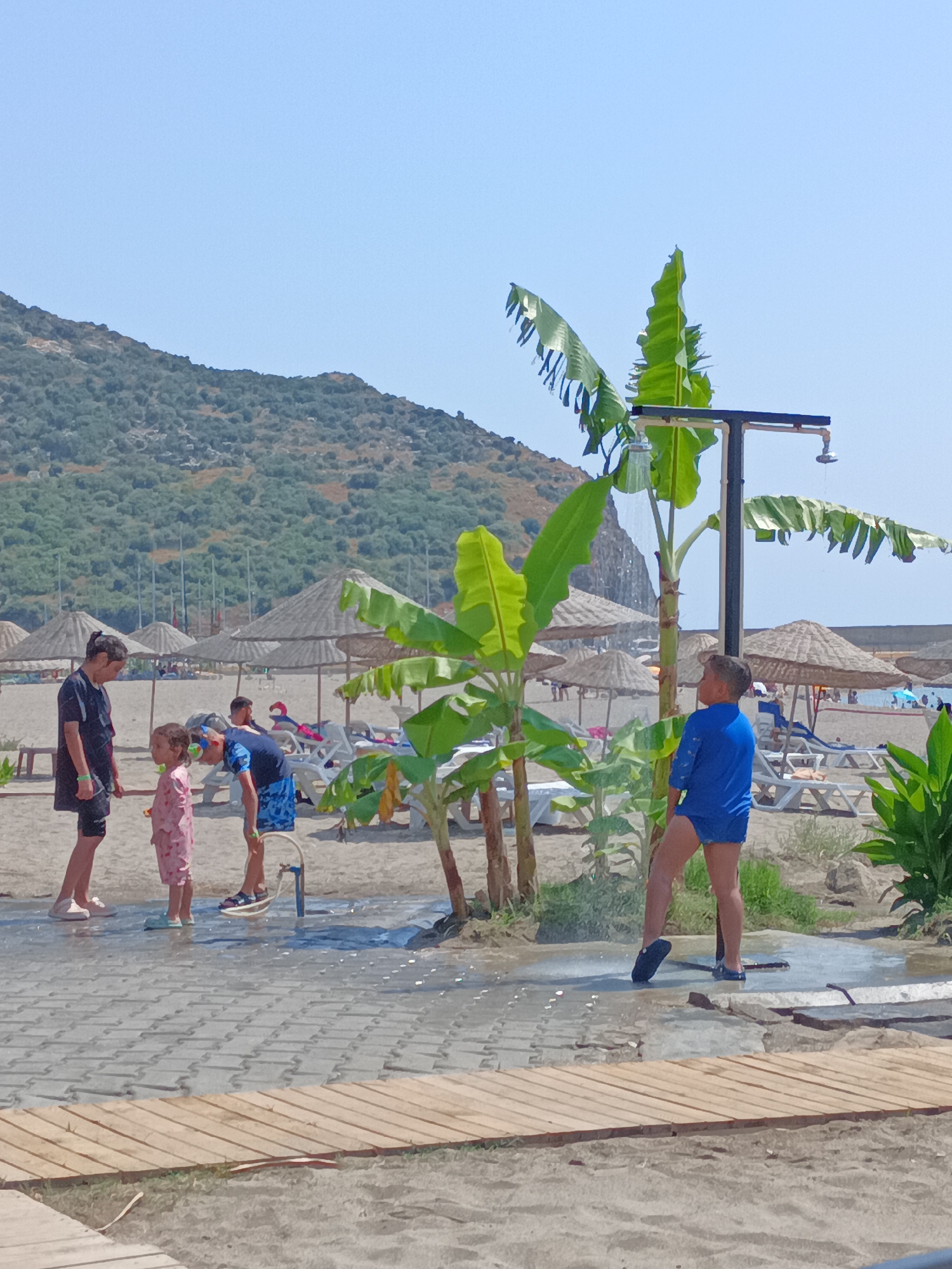In Gazipaşa, Turkey, on a sunny day at the beach, several individuals are visible near an outdoor shower. Two children, a girl in pink pajamas and a boy in blue shorts and a darker blue t-shirt, stand near the shower. A third child, a boy wearing a blue swimsuit, is actively using the shower, with water spraying from above. Nearby, a man in a black t-shirt and shorts stands with his back to the camera, facing the children and the shower. Further in the background, beach umbrellas and lounge chairs are set up on the sand. The landscape includes a tree with large leaves, possibly a banana tree, and a hill covered in vegetation. The ground in the foreground is a tiled pathway that appears wet from the shower.